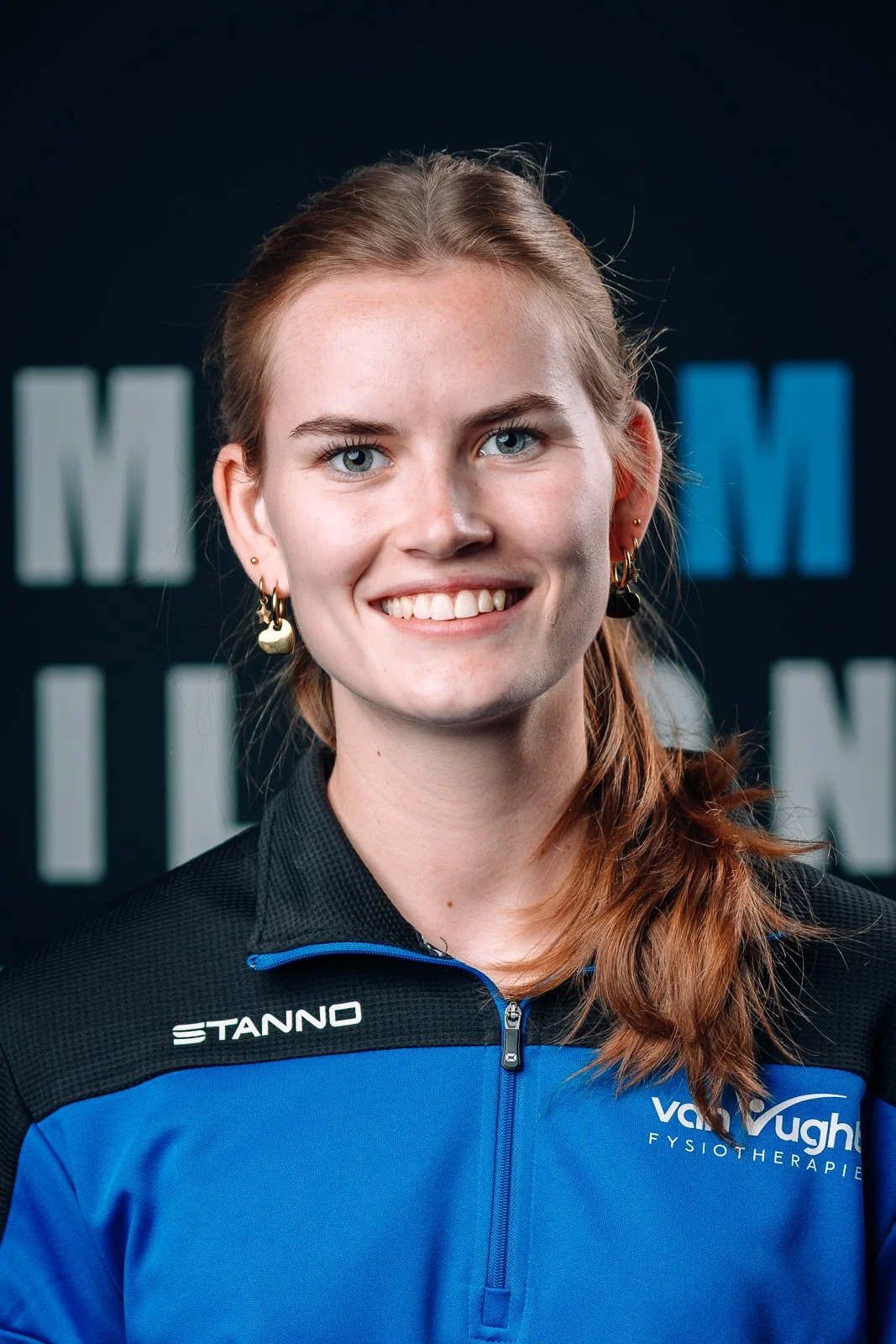 Een jonge vrouw met lang, roodbruin haar, blauwe ogen, die een sportjasje draagt met het logo 'VogVaugh Fysiotherapie' en eeretattoos in haar oren, lachend voor een donkere achtergrond met grote letters.