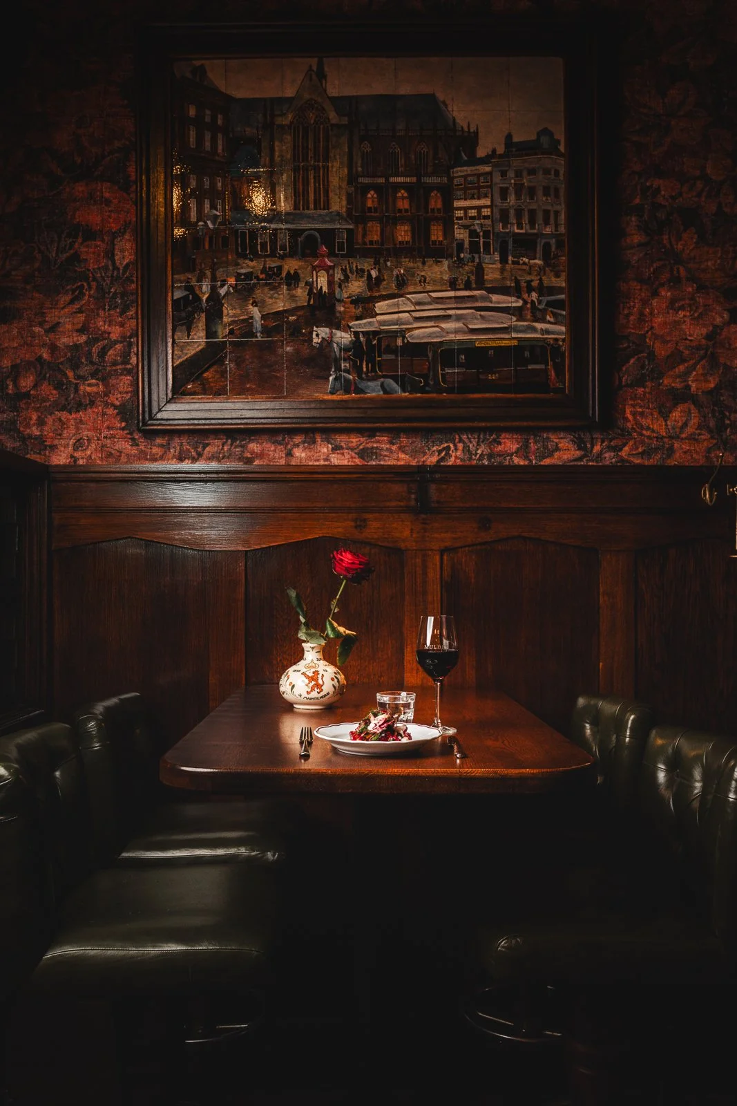 Een donker, knus restaurant met een houten tafel, een rode roos in een vaas, een glas rode wijn, een bord met voedsel, en een schilderij van een stad met een kathedraal op de muur.