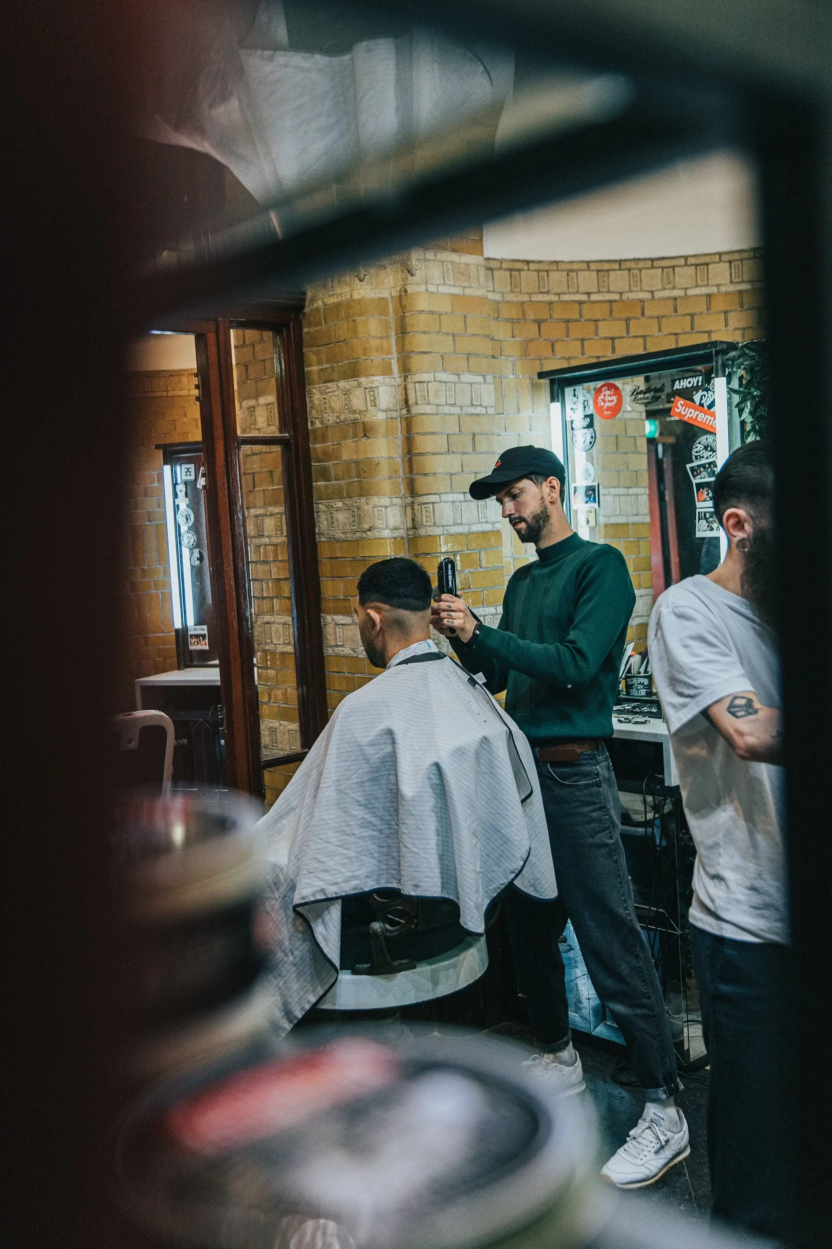 Man met een cap en een groene trui die een paar jongens achter een kappersstoel in een kapsalon knipt, tegen een muur van bakstenen met een spiegel met stickers.