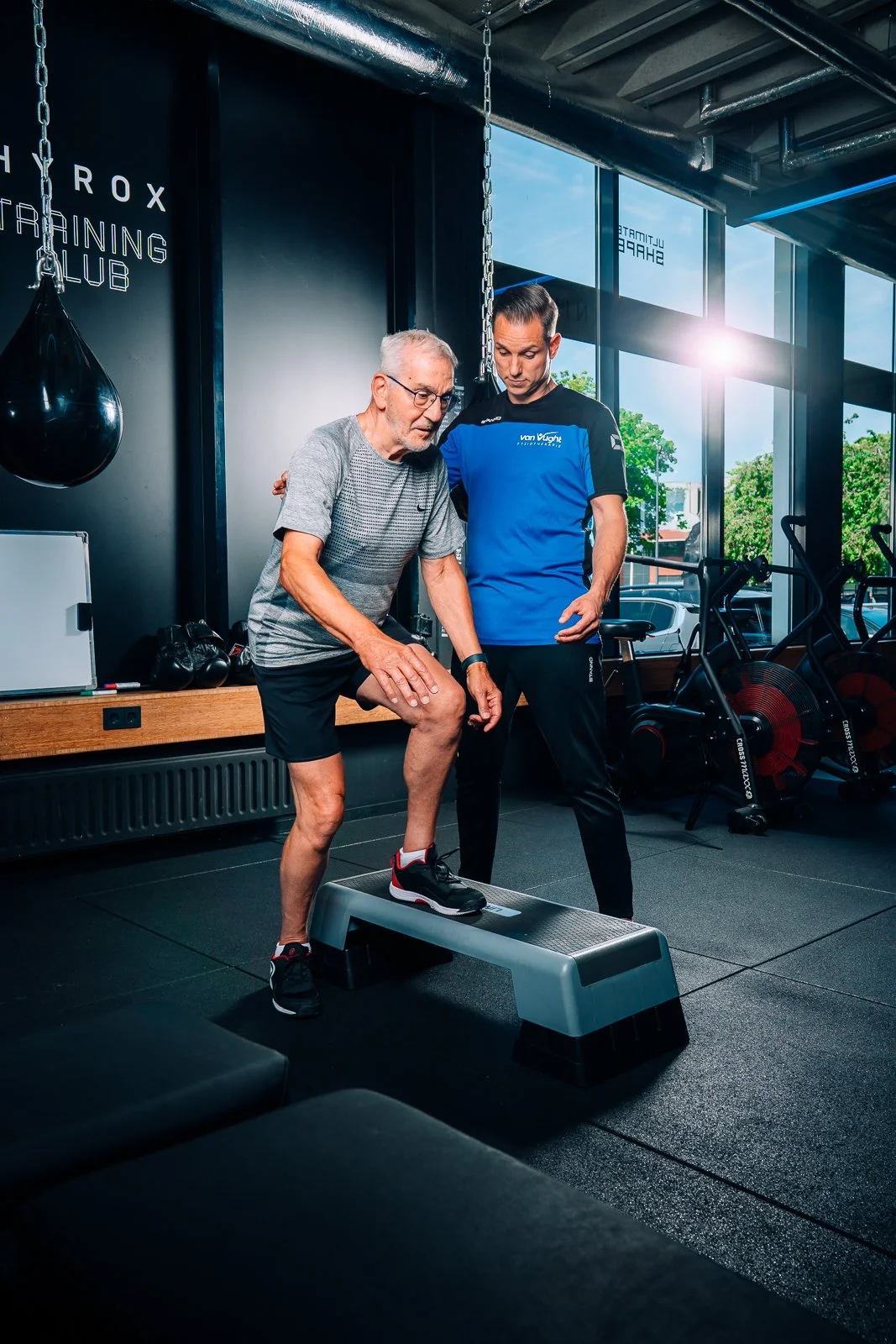 Personal trainer helpt oudere man met hardloopoefeningen op een step in een sportschool met grote ramen en fitnessapparatuur op de achtergrond.