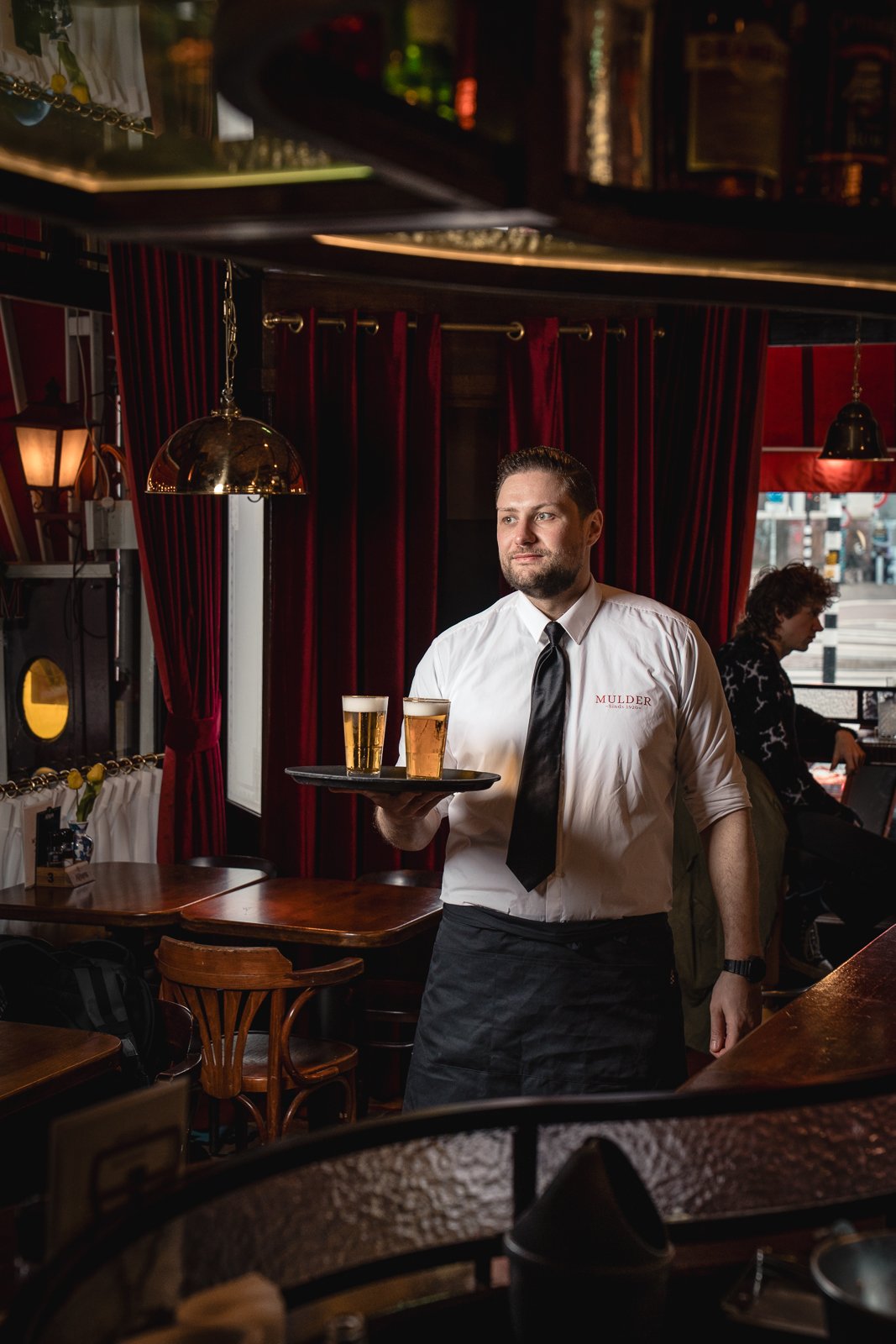 Kelner in een witte blouse, zwarte stropdas en zwarte schort, die twee bieren serveert in een gezellig restaurant met rode gordijnen en houten meubels.
