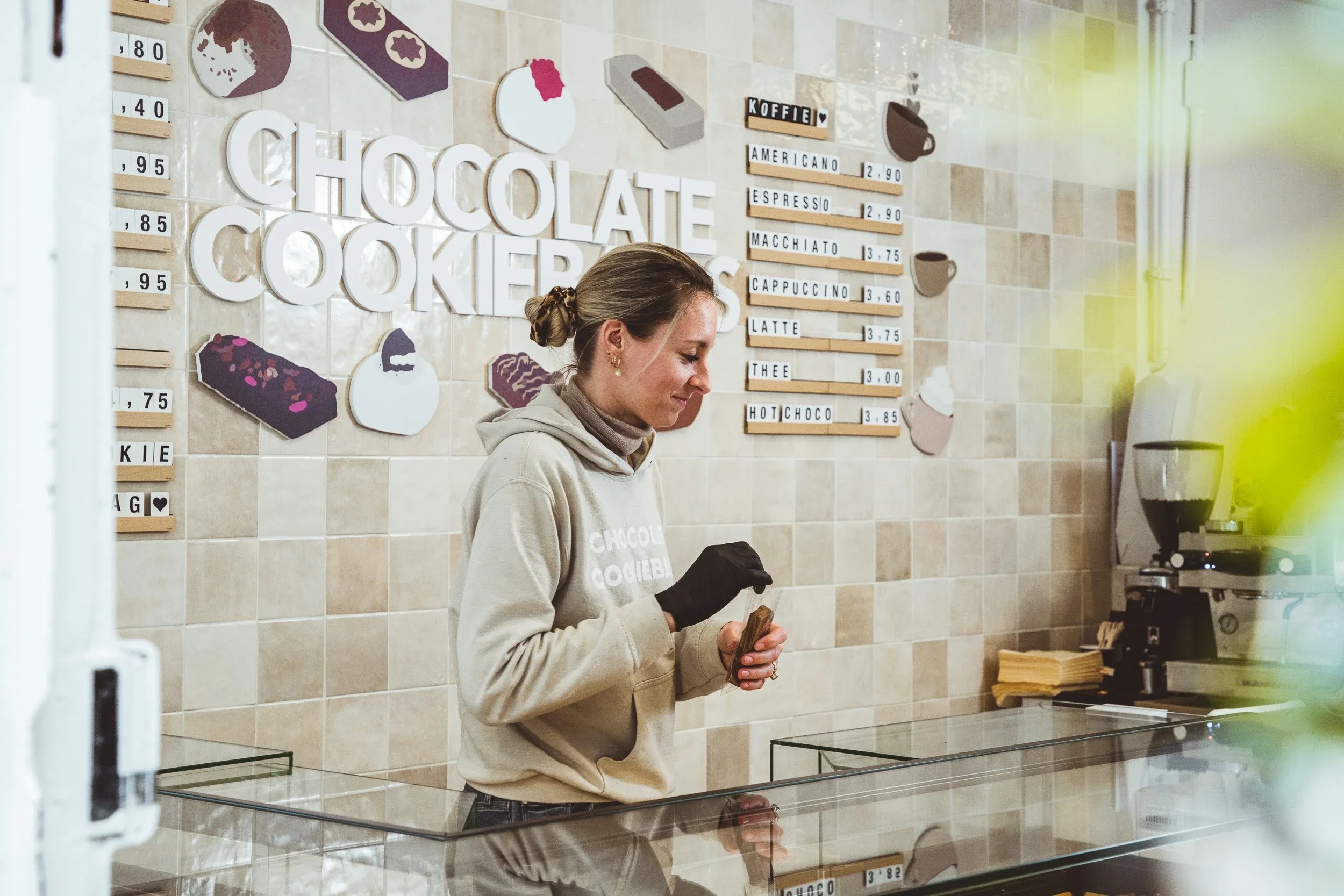 Een vrouw met een beige hoodie en een paar oorbellen, die binnen in een koffiezaak staat, terwijl ze haar telefoon vastpakt.
