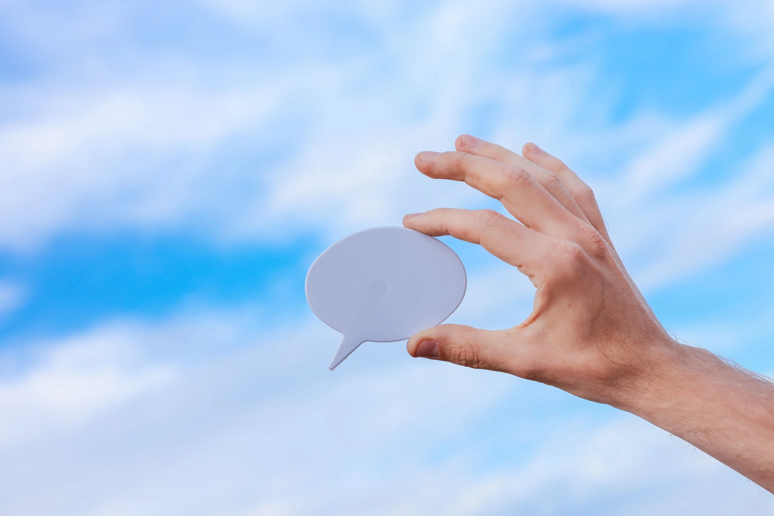 A person's hand holding a white speech bubble cutout against a blue sky with some clouds.