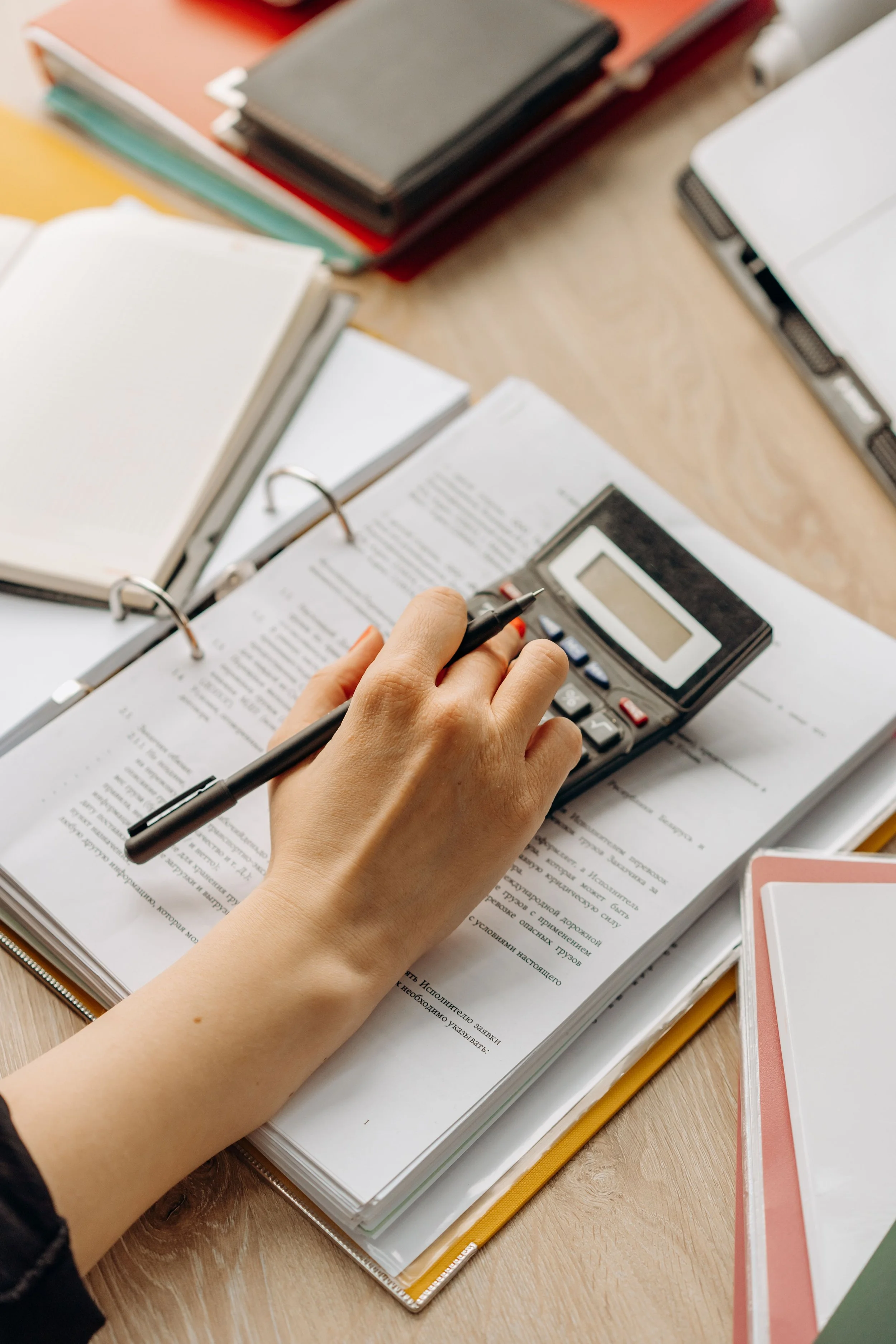 A person using a calculator, surrounded by notebooks, papers with text, and colored sticky notes on a wooden desk, possibly studying or working.
