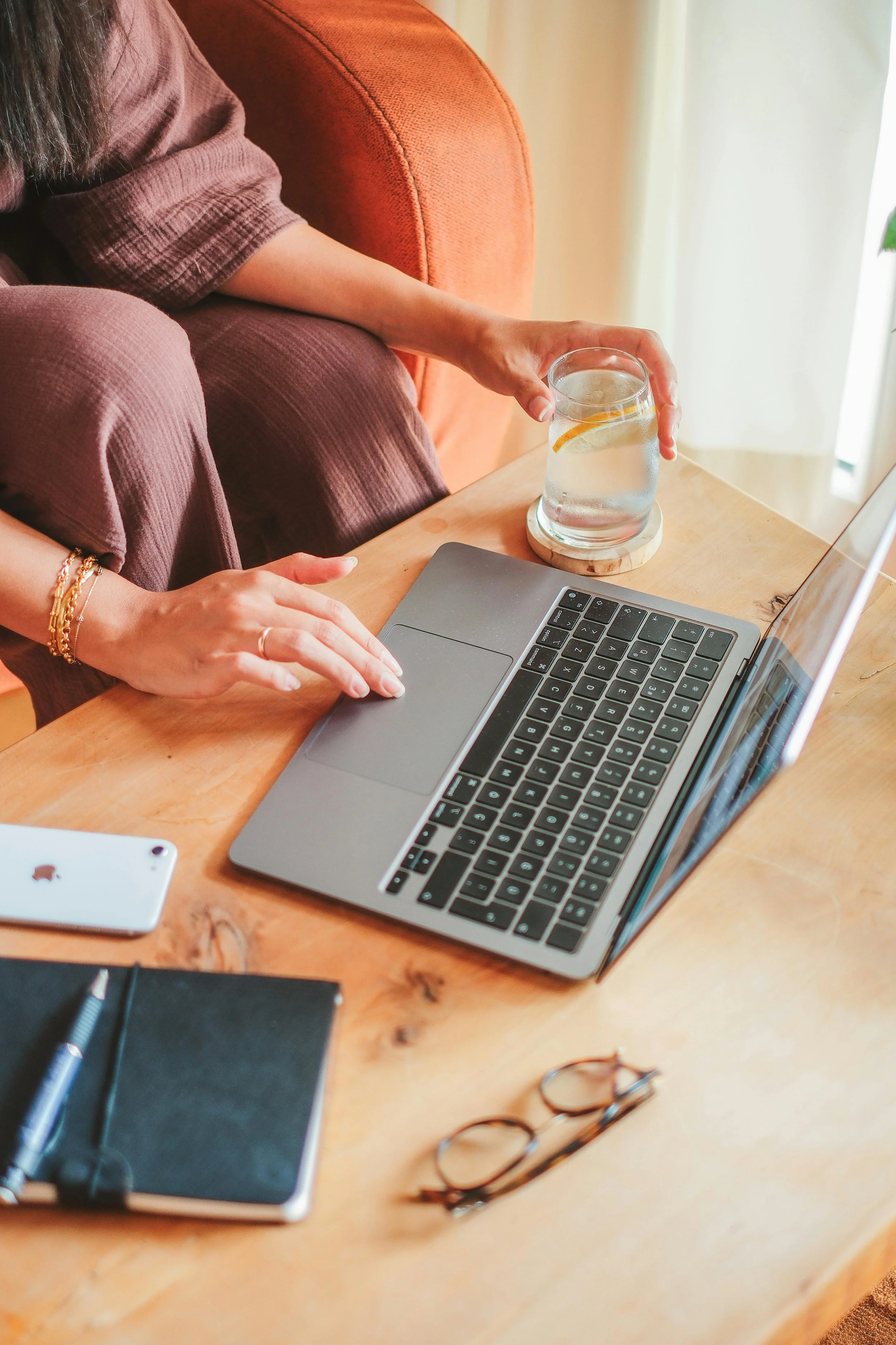 A person sitting at a wooden table with a laptop, a glass of water with lemon, and various stationery items, including a pair of glasses, a notebook, and a pen.