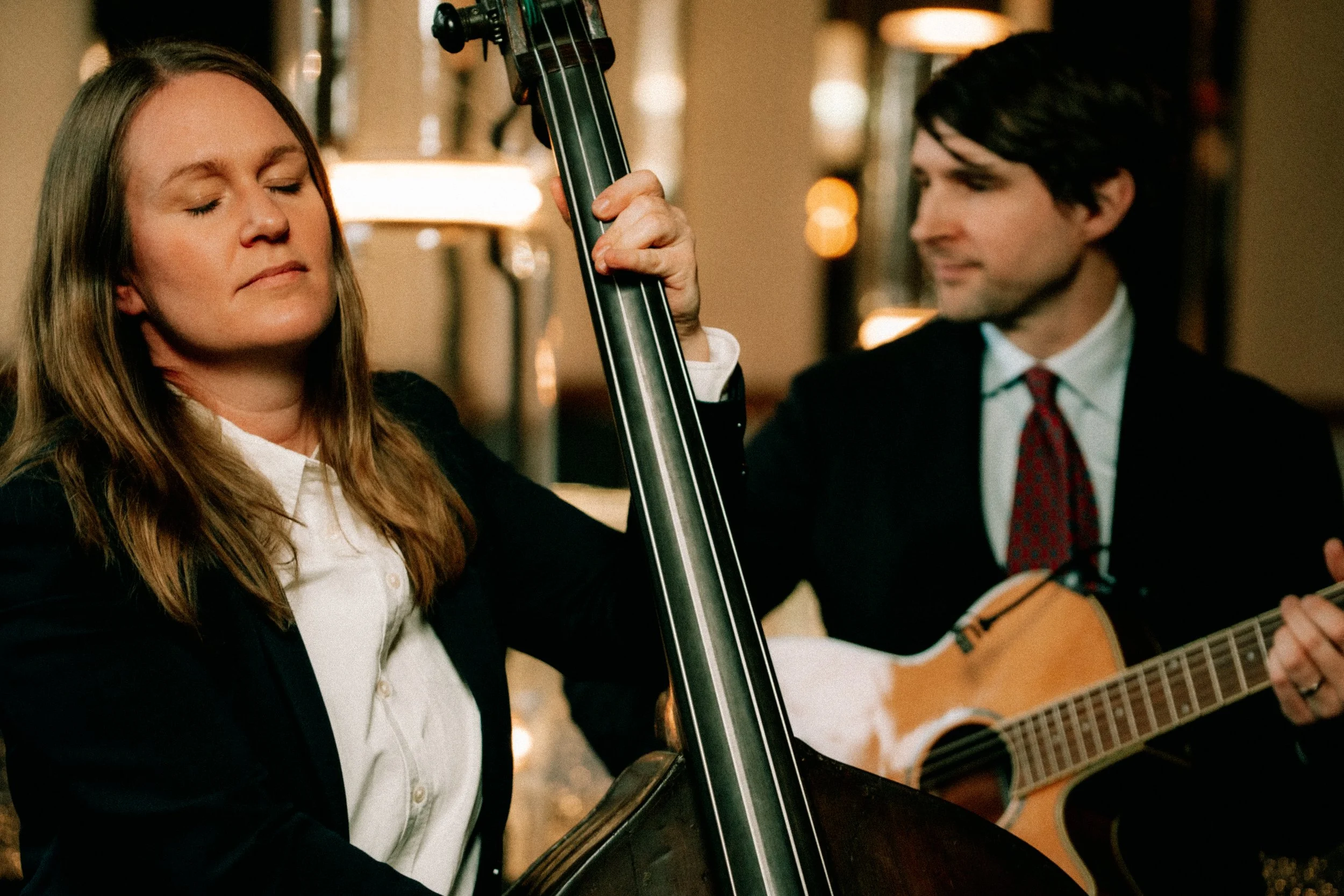 Två personer, en kvinna med blont hår och en man med mörkt hår, spelar gitarr och står nära varandra innomhus, på en tillställning eller i ett cafe.