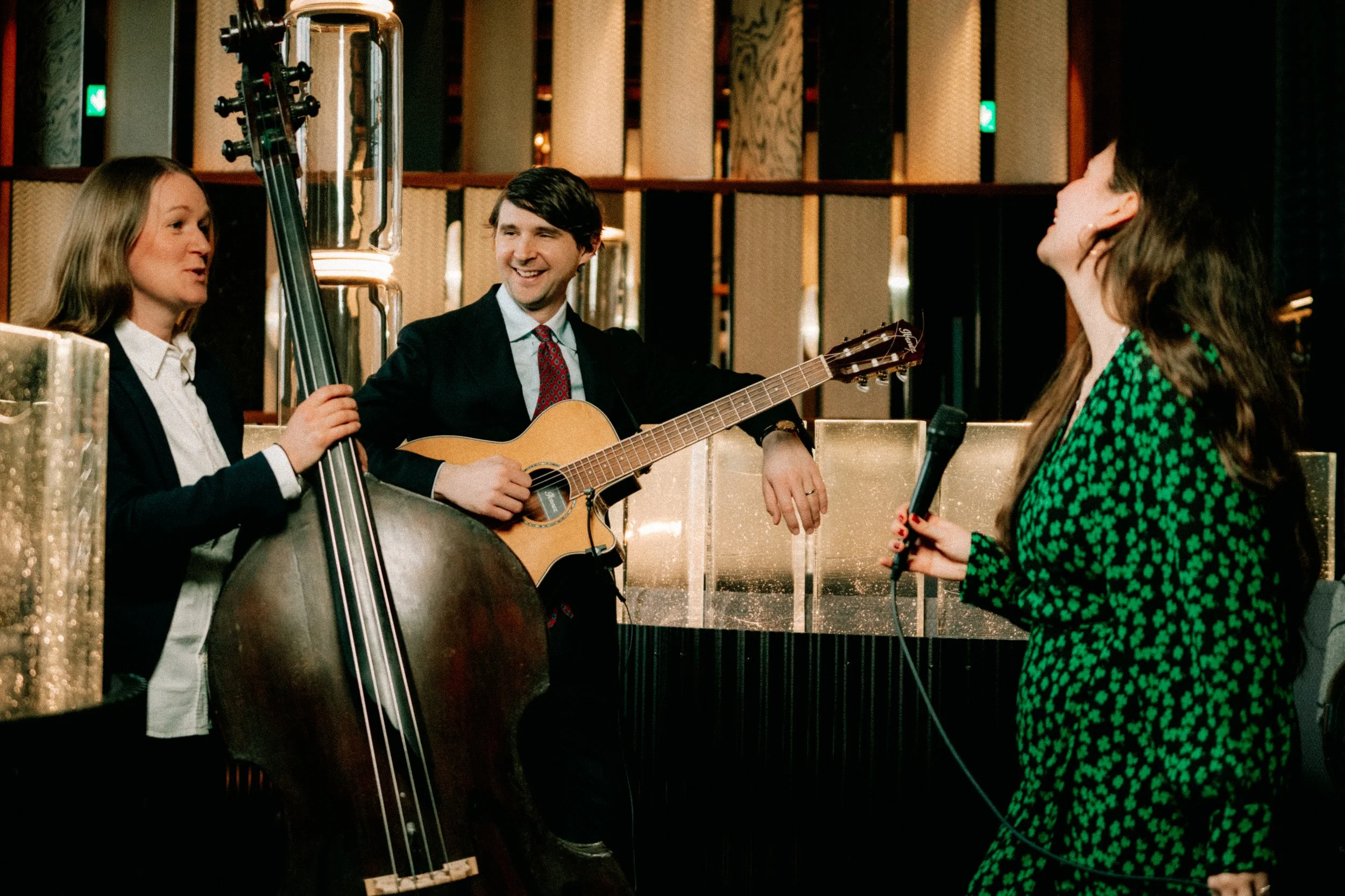 Tre personer, två kvinnor och en man, som alla är glada, samtalar och leker musik tillsammans i en elegant inomhusmiljö. Mannen spelar gitarr, en kvinna håller en mikrofon, och den andra kvinnan lyssnar och deltar i samtalet.
