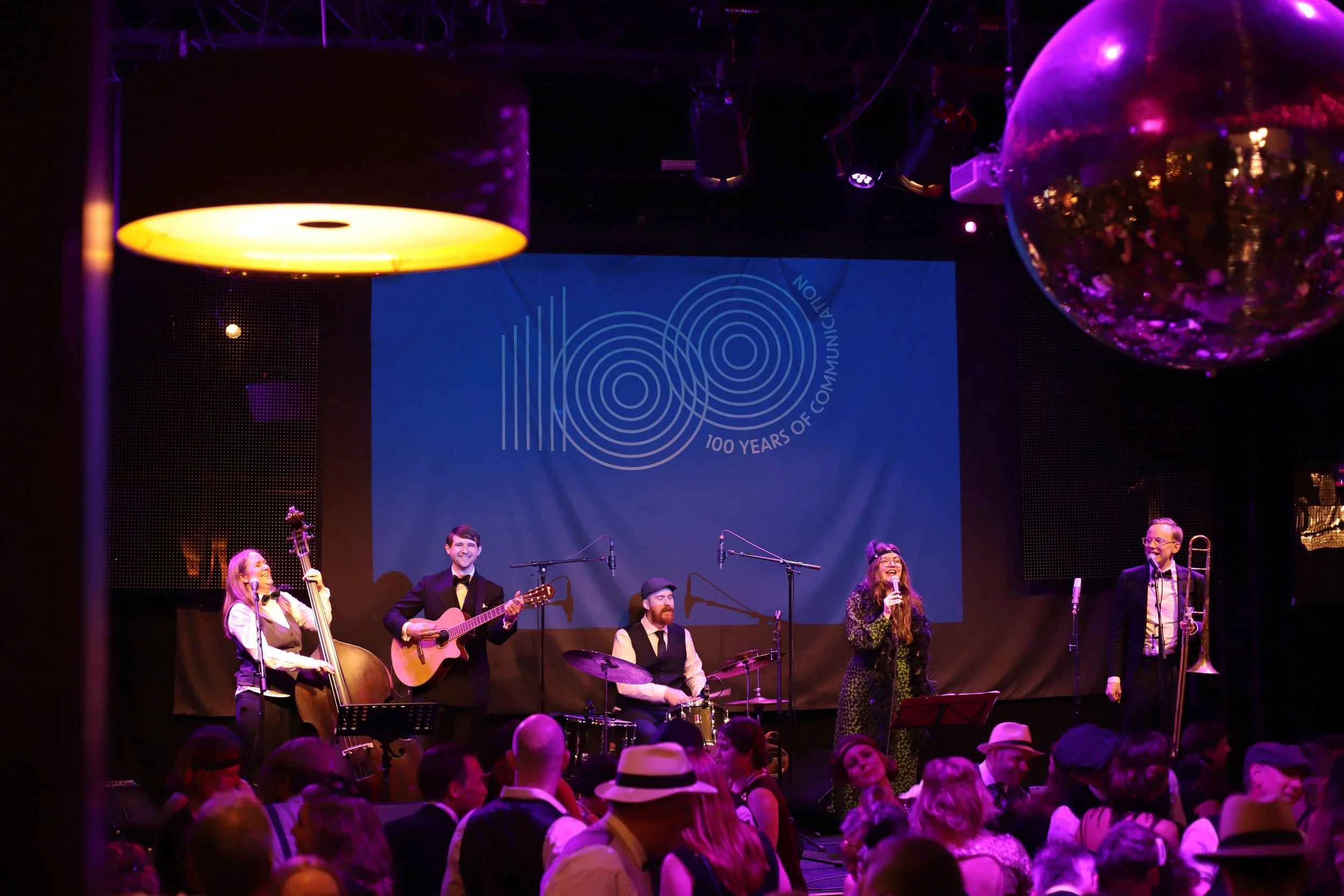 Musiker som spelar på scenen under en konsert med en mörk bakgrund, en stor skärm med text '100 Years of Communication', festliga dekorationer och publik i förgrunden.