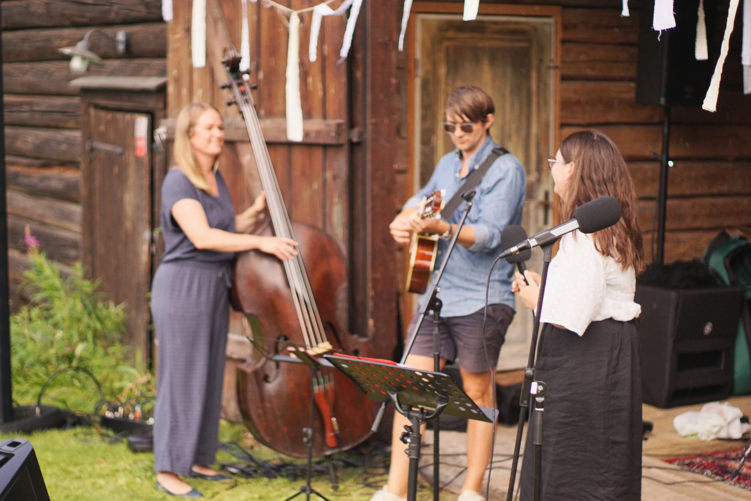 Tre musiker som spelar instrument, en med kontrabas, en med gitarr och en med mikrofon, under ett utomhusframträdande framför en gammal trävägg.