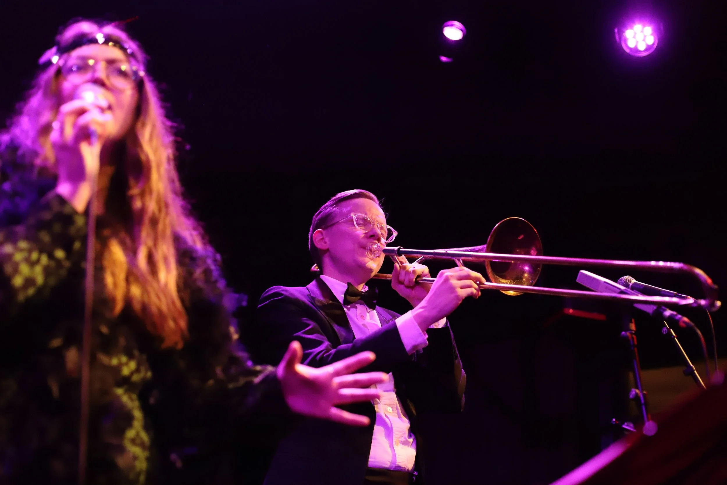 Två musiker, en kvinna och en man, spelar live på scen under en konsert. Kvinnan sitter till vänster, har långt, lockigt hår och bär glasögon, medan mannen i mitten spelar trombon och bär en svart smoking och glasögon.