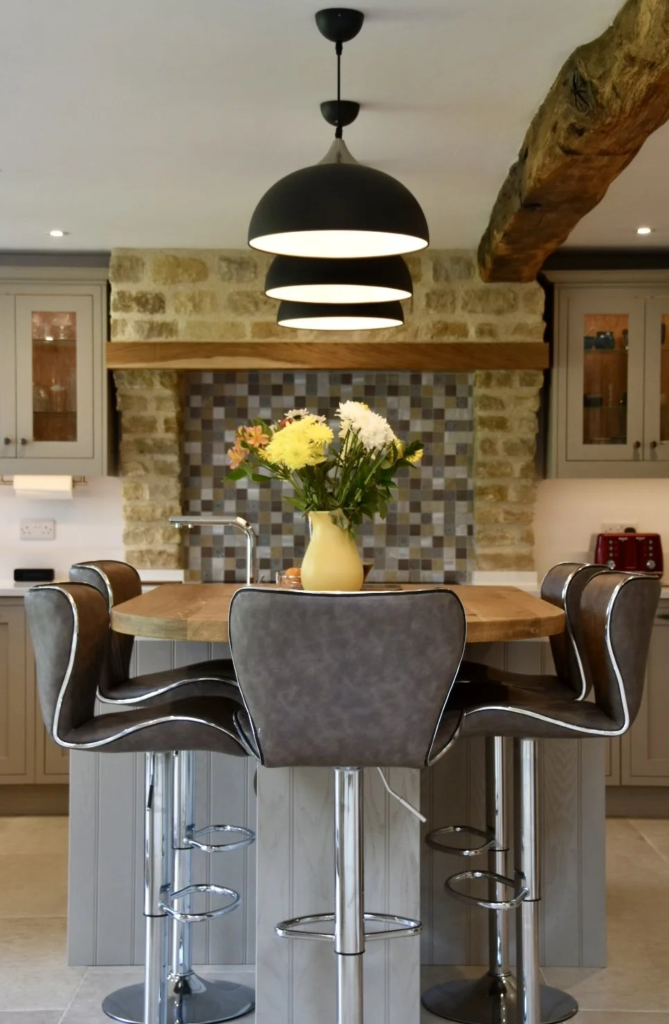 Kitchen with a round wooden island, black bar stools, a vase with flowers, pendant lights, and a brick wall background.
