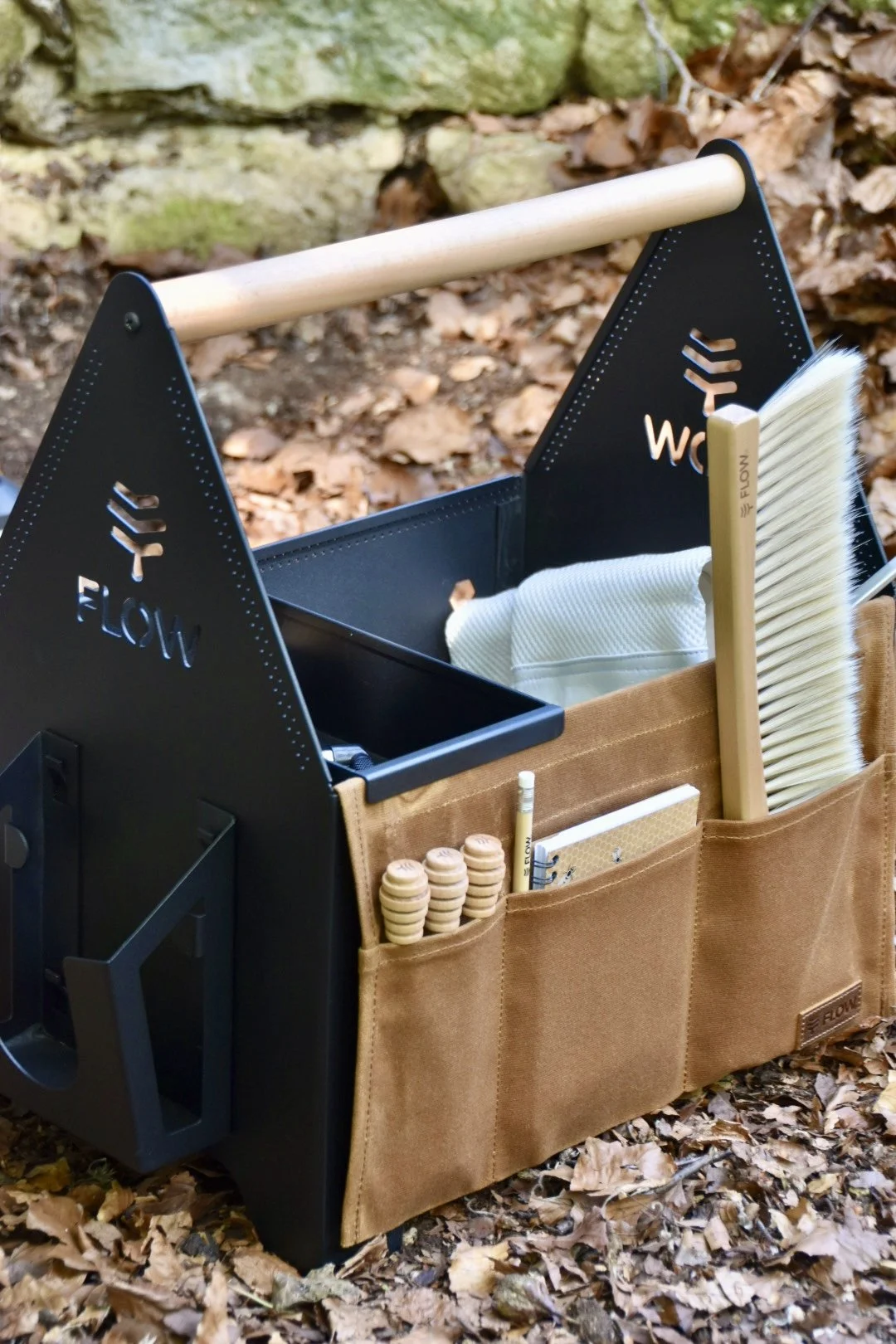 A black and tan outdoor work station with a wooden handle, brushes, notebook, and small tools, situated on fallen leaves in a natural setting.