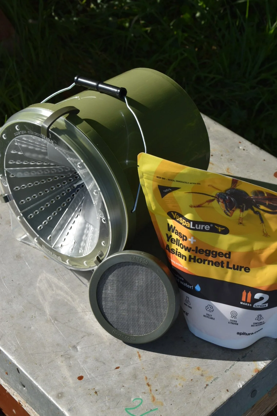 A metal wasp trap with a yellow and black bag labeled "VespaLure" indicating it's a wasp lure, an empty bait pod, and a gray round speaker or filter, all placed on a weathered table outdoors with grass in the background.