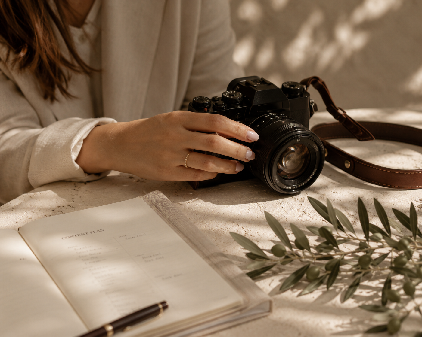 A woman in a beige blazer holding a vintage black camera with a brown strap on a textured surface with an open planner, a black pen, and green foliage.