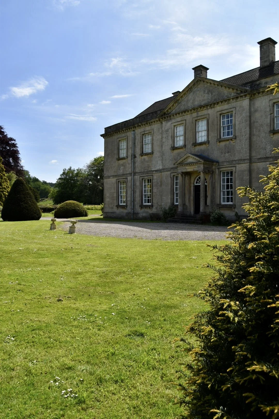 A large historic mansion with a stone exterior and multiple windows is set in a well-manicured lawn on a sunny day, with a clear blue sky above.