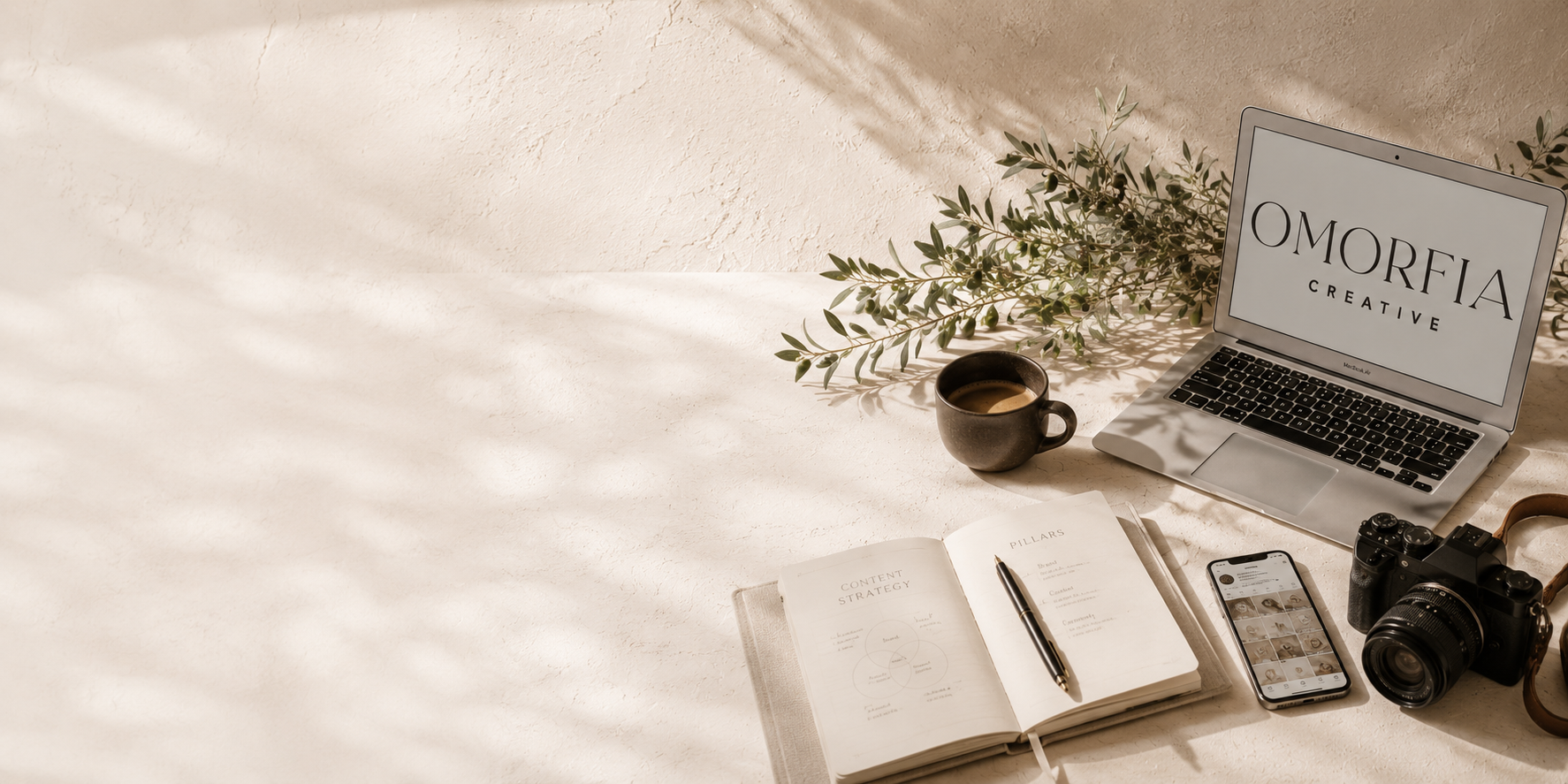 A workspace with a laptop displaying 'OMORFIA CREATIVE', an open notebook with a pen, a smartphone showing social media images, a camera, a cup of coffee, and a sprig of greenery on a light-colored textured surface.