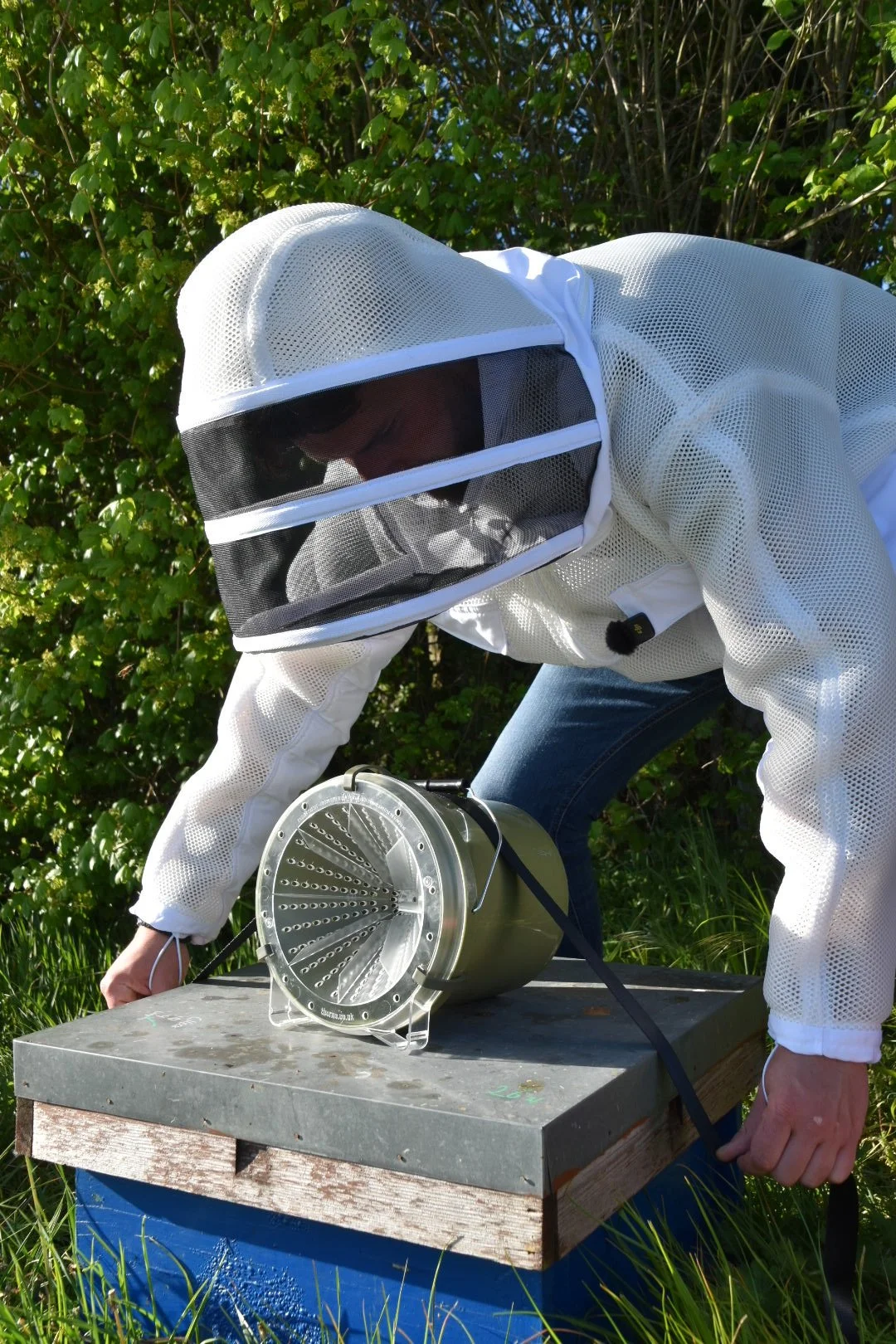 Person wearing a white protective beekeeping suit and a veil inspecting a honey extractor in an outdoor setting with green foliage.