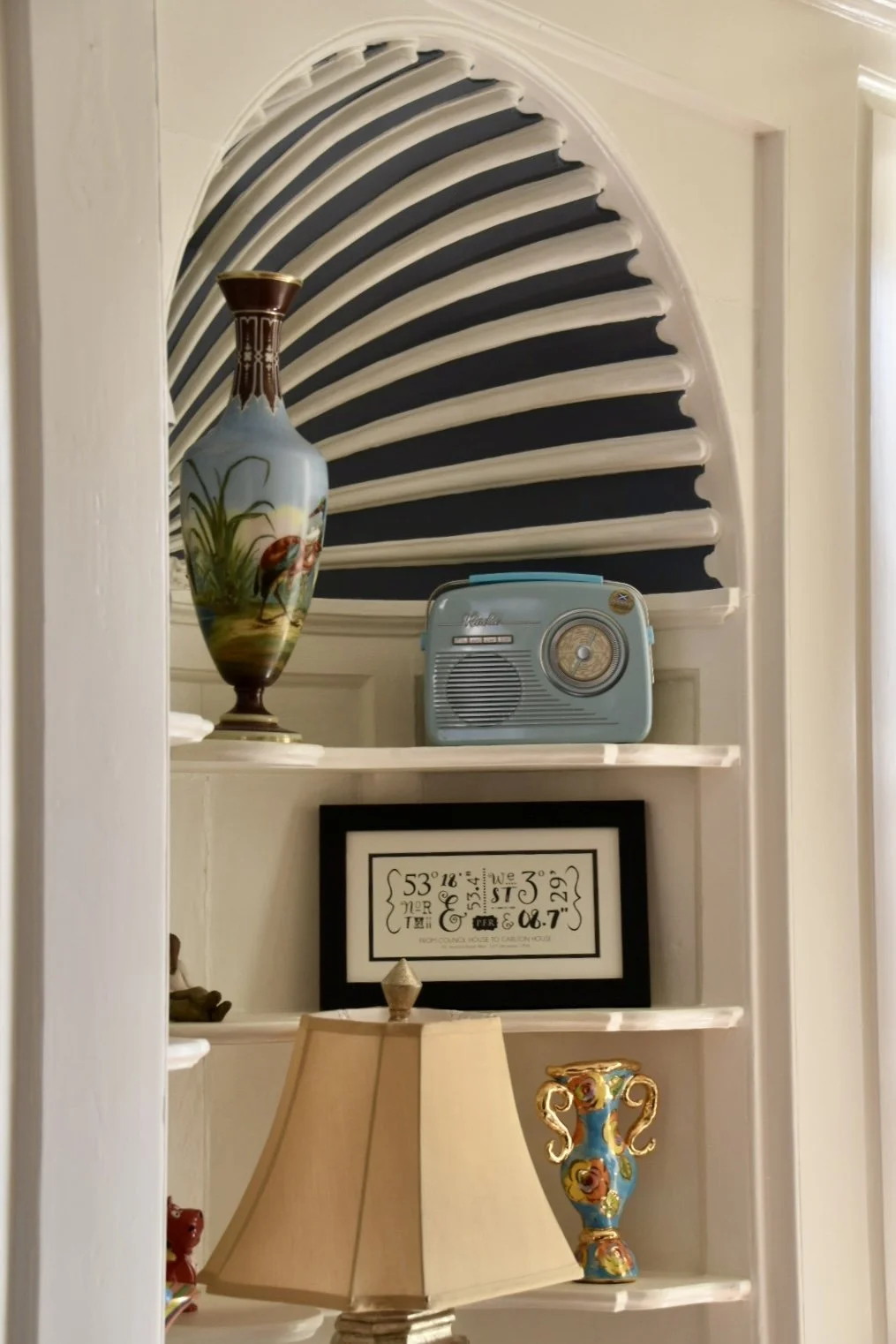 A white built-in wall shelf with decorative items including a large painted vase, a vintage blue radio, a framed art print, a small lamp, and a colorful vase. There is an arched window with black and white striped blinds behind the shelf.