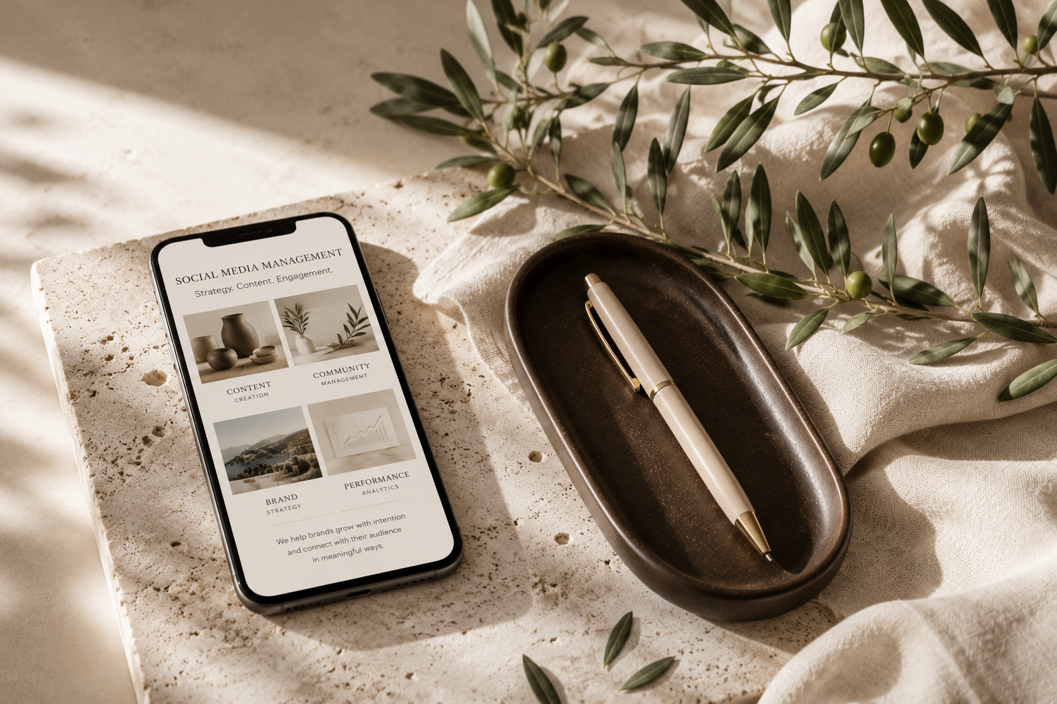 Smartphone displaying social media management content, a white pen in a brown ceramic dish, green olive branch on a textured beige surface with soft lighting.