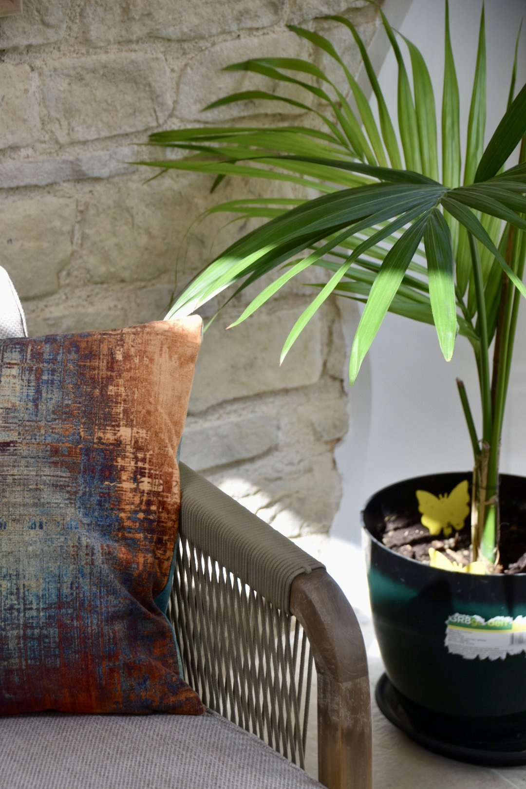 A corner of a room with a gray cushioned chair, a colorful textured pillow, a large green potted plant with long leaves, and a stone wall in the background.