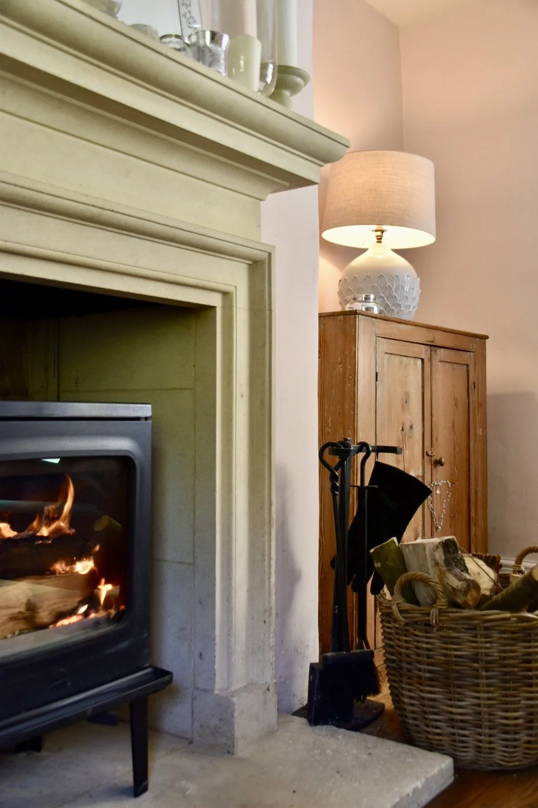 Living room with fireplace, wooden cabinet, lamp, basket of firewood, fireplace tools, and a basket of firewood.