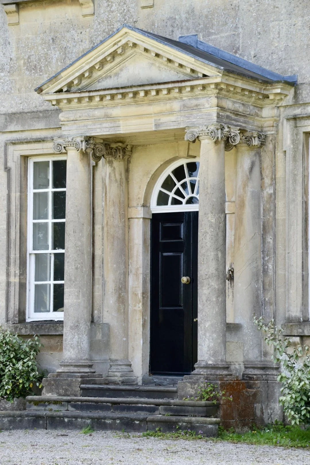 An old stone building with a black front door, two classical columns, and a triangular pediment above the entrance.