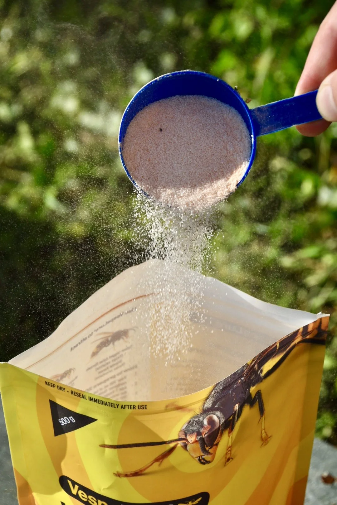 A hand holding a blue scoop of pink insecticide powder over an open yellow bag with a picture of a wasp on it, outdoors with green foliage in the background.