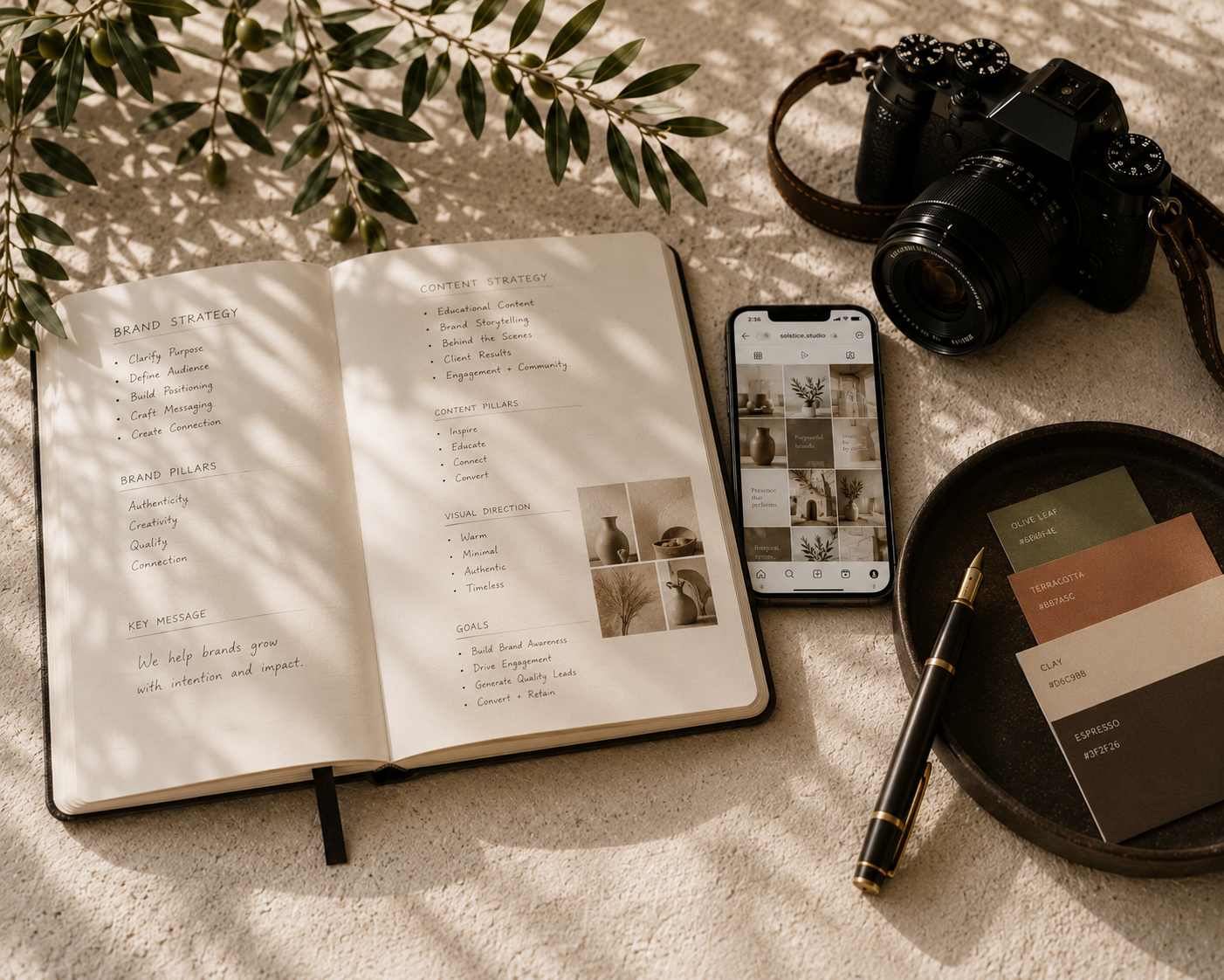 Photography workspace with an open notebook, a smartphone displaying a photo grid, a digital camera, a tray with color swatches, and a pen, all on a light textured surface with shadows of leaves.