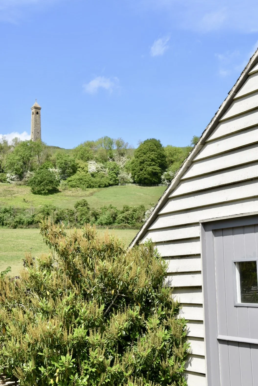 A house with light gray siding and a small window, surrounded by green bushes, with a lush green hillside, trees, and a tall tower on the hill in the background under a blue sky.