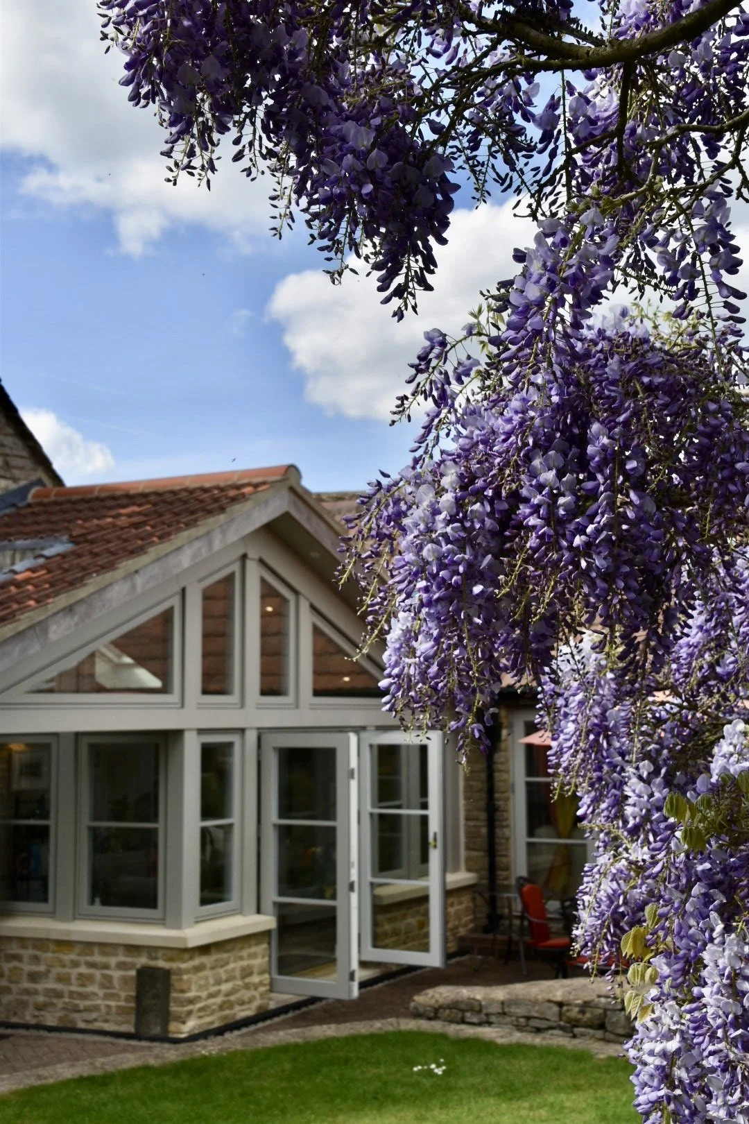 A house with large windows and a door, surrounded by purple flowering wisteria vines hanging from a tree, under a partly cloudy sky.