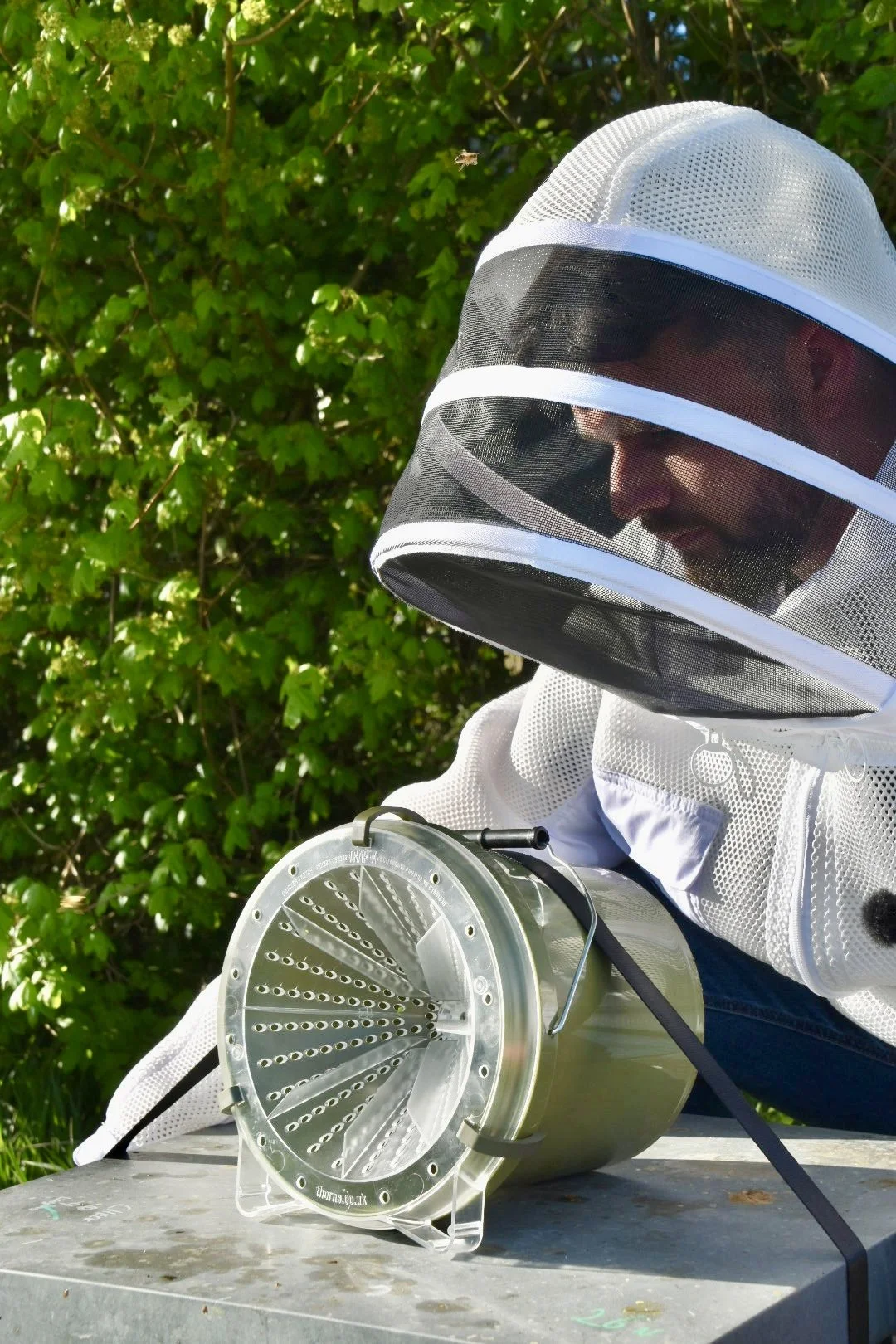 Person in protective hat and mesh face covering holding a honey extractors on an outdoor table with green foliage in the background.