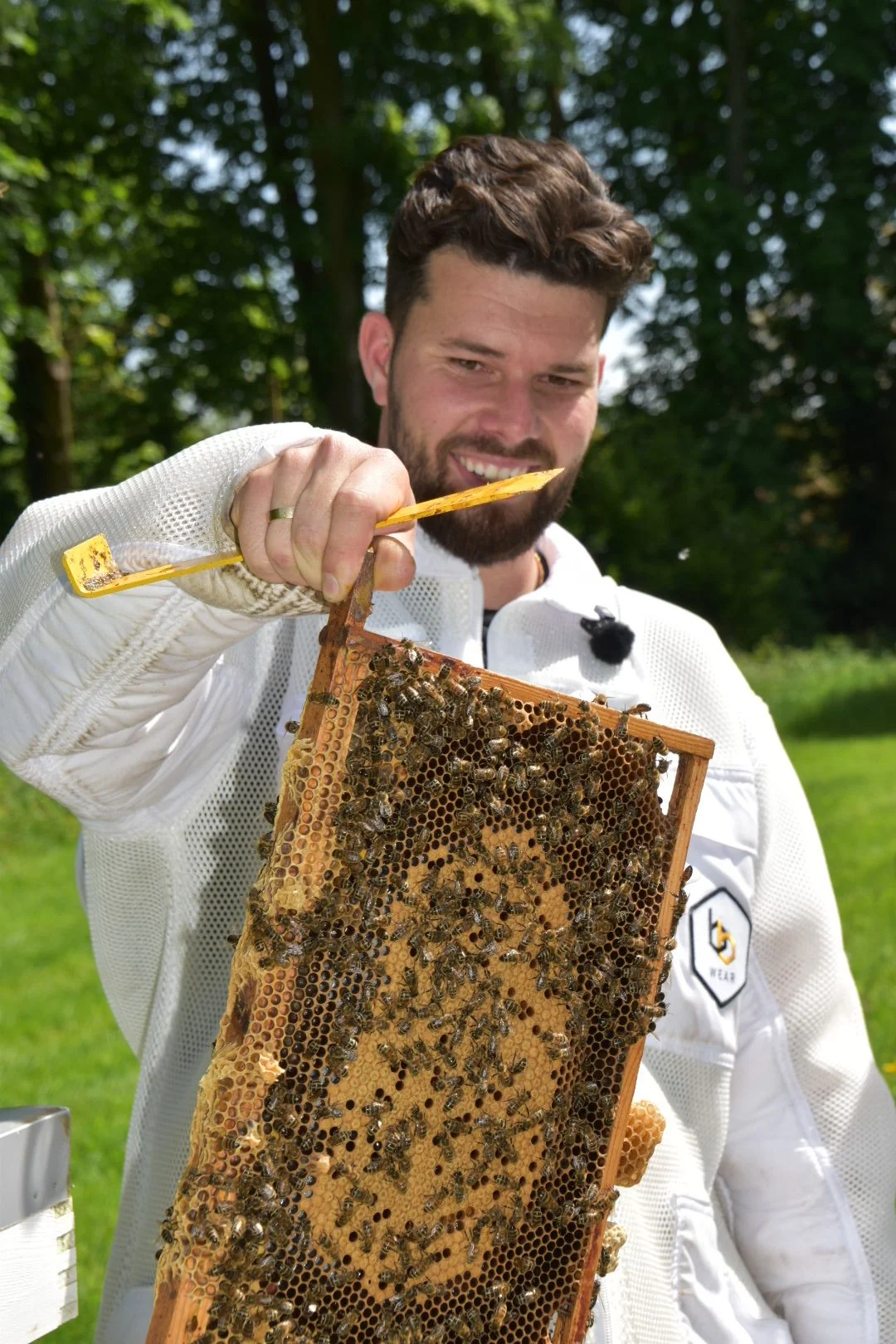 A man with a beard and brown hair inspecting a honeycomb frame covered with bees outdoors, with trees and grass in the background.