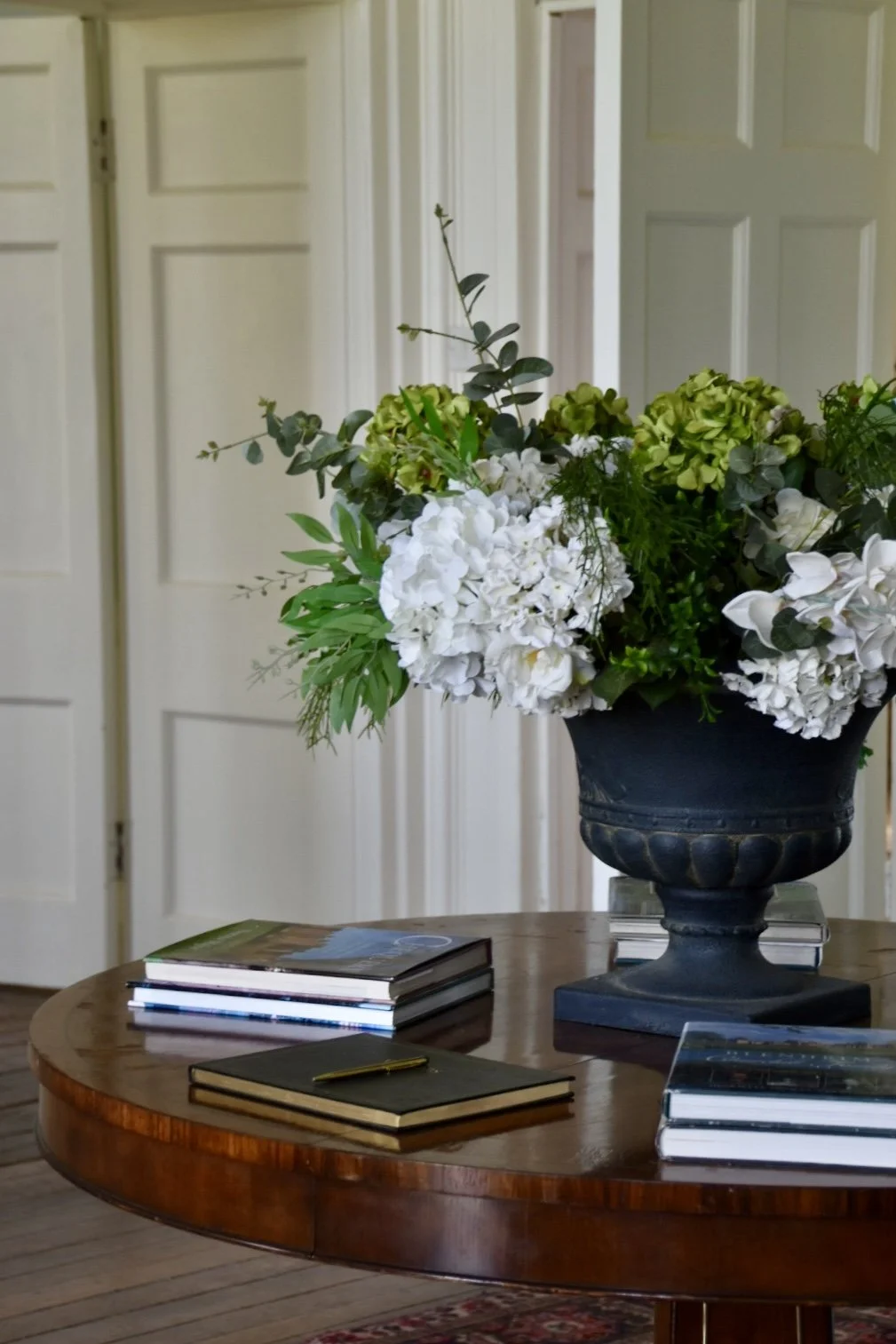 A round wooden table with a large black vase containing white and green flowers, placed on top of a few books with a black notebook and gold pen, in a room with white paneled doors in the background.