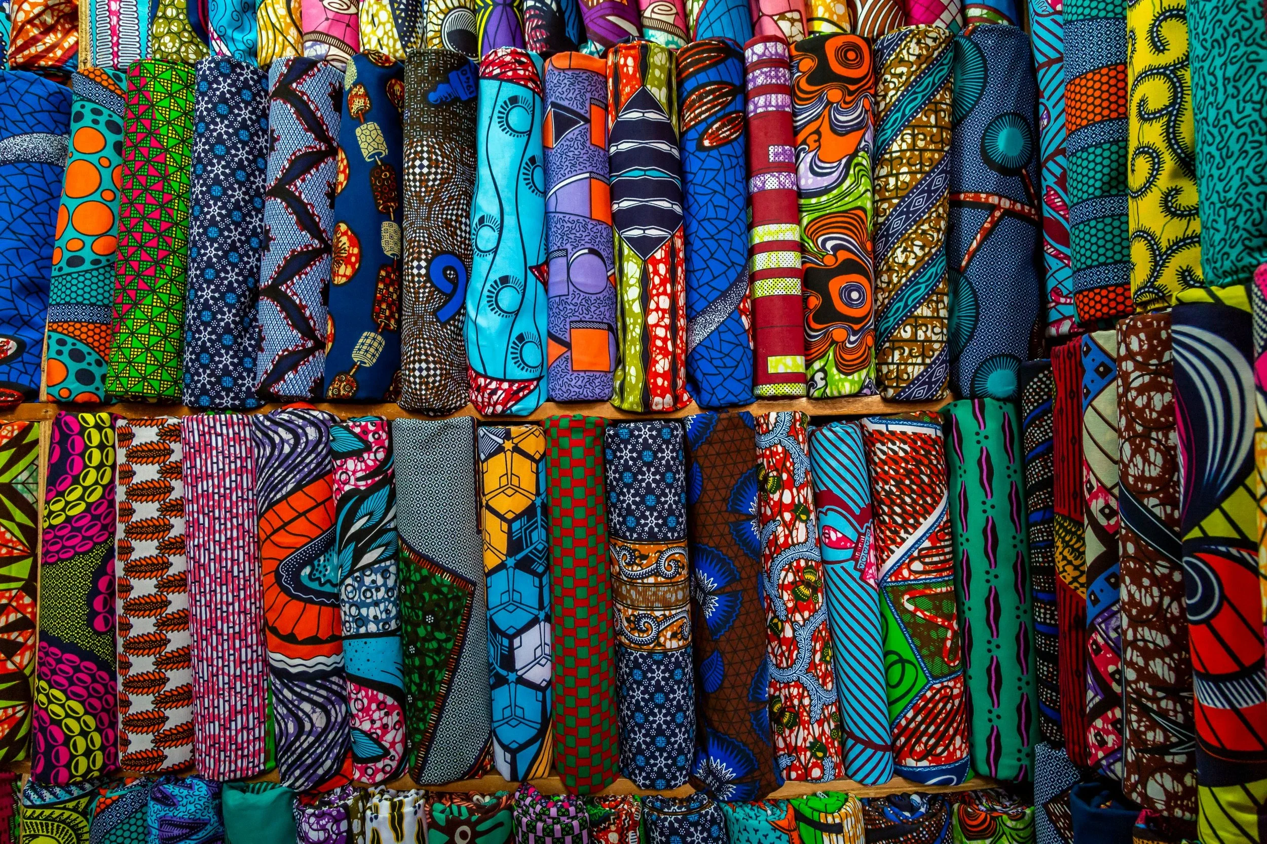 A display of rolls of colorful fabric with various vibrant African patterns arranged vertically on a shelf.
