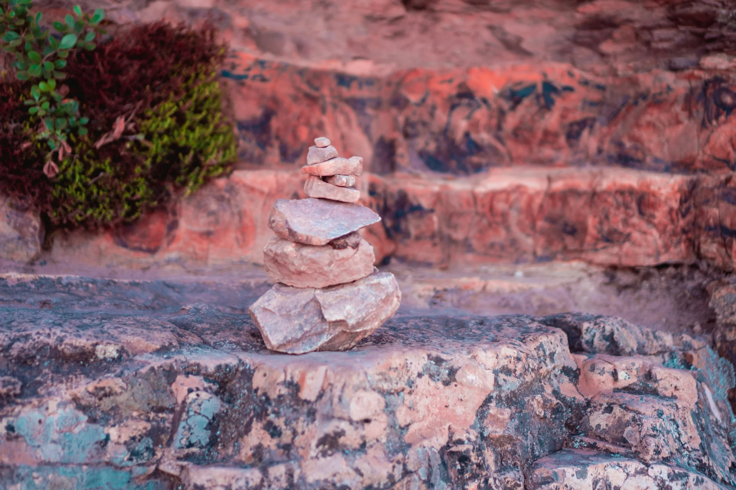 A small rock cairn with several flat stones stacked on top of each other on a rocky surface, with a green bush and red rock background.