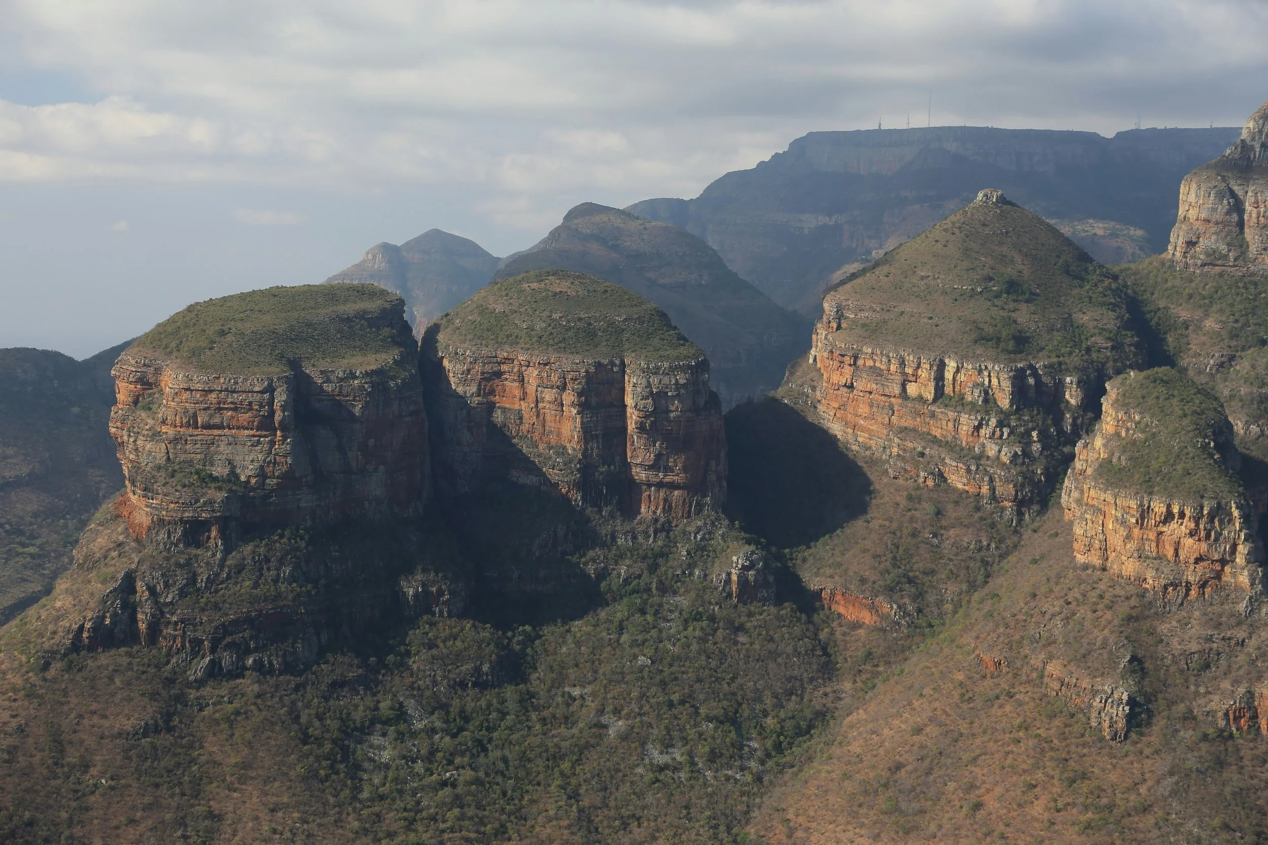 A scenic view of a South African mountain range with layered rocky formations, green vegetation, and a cloudy sky overhead.
