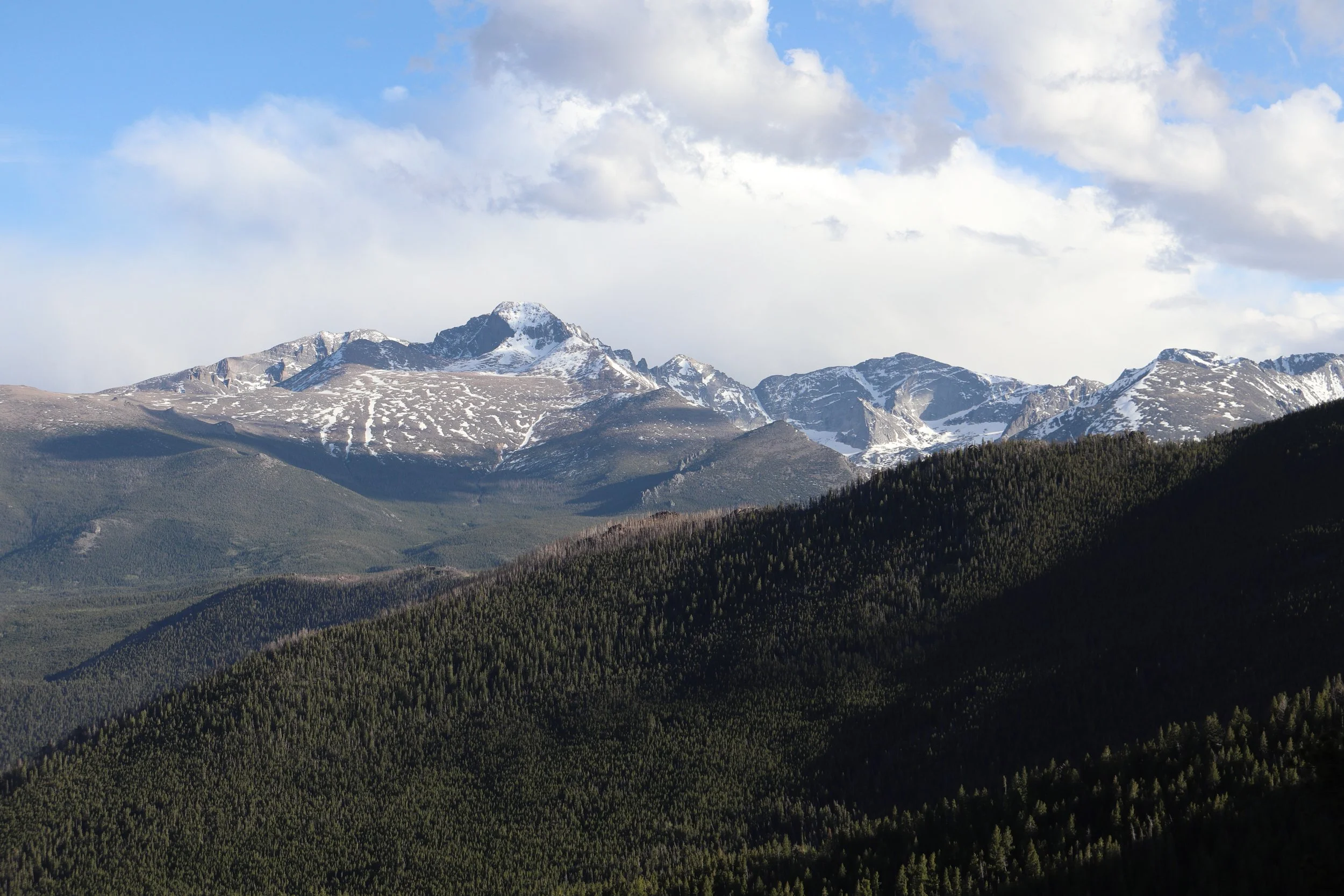 Snow-capped mountains under a partly cloudy sky with layers of green forest in the foreground.
