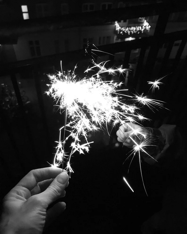 Black and white photo of two hands holding lit sparklers at night on a balcony, bright sparks radiating against a dark background with silhouetted buildings.
