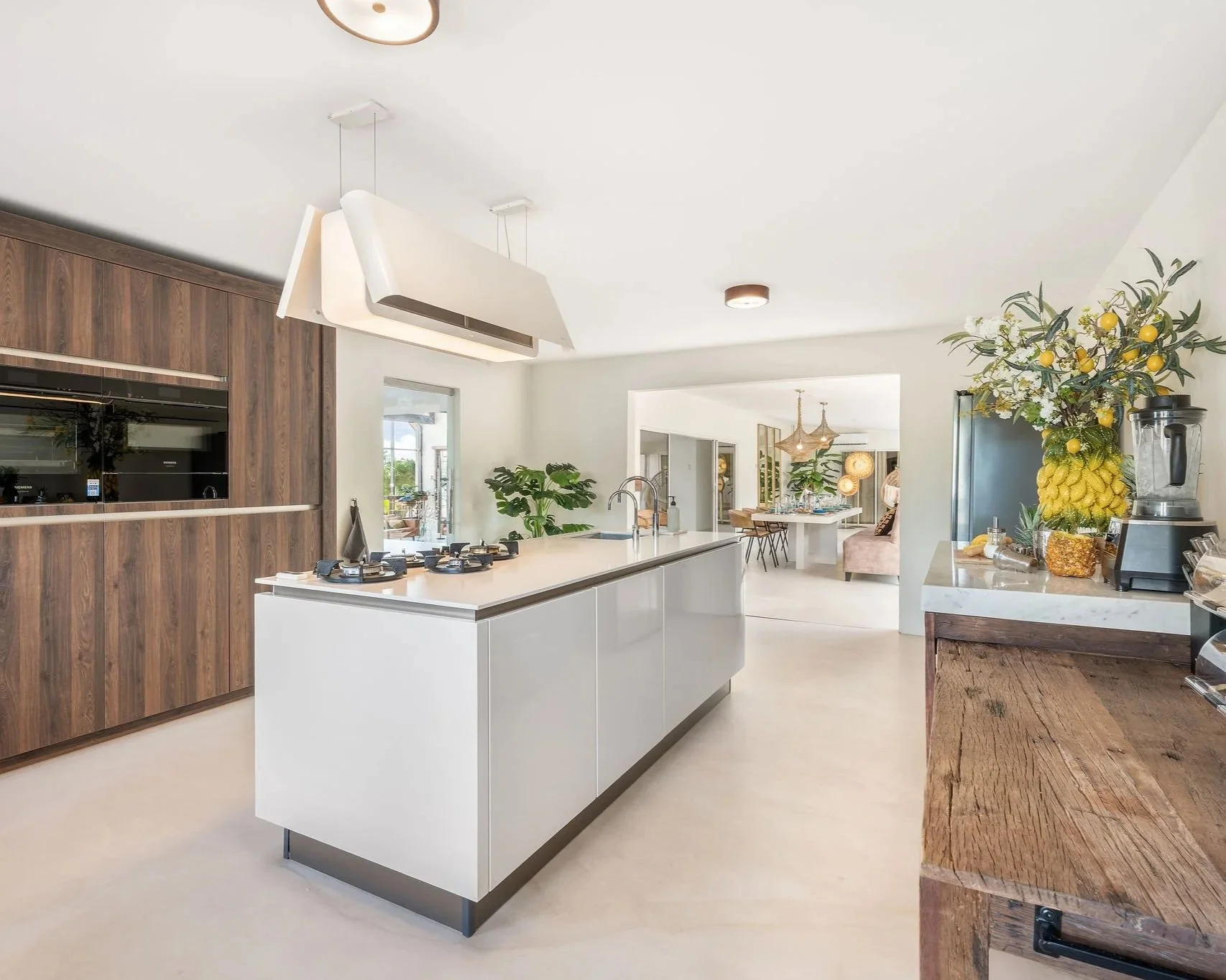Open-concept modern kitchen with white island, wooden cabinetry, potted plants, and a view into adjacent dining area with hanging light fixtures and a sitting area with a pink chair.