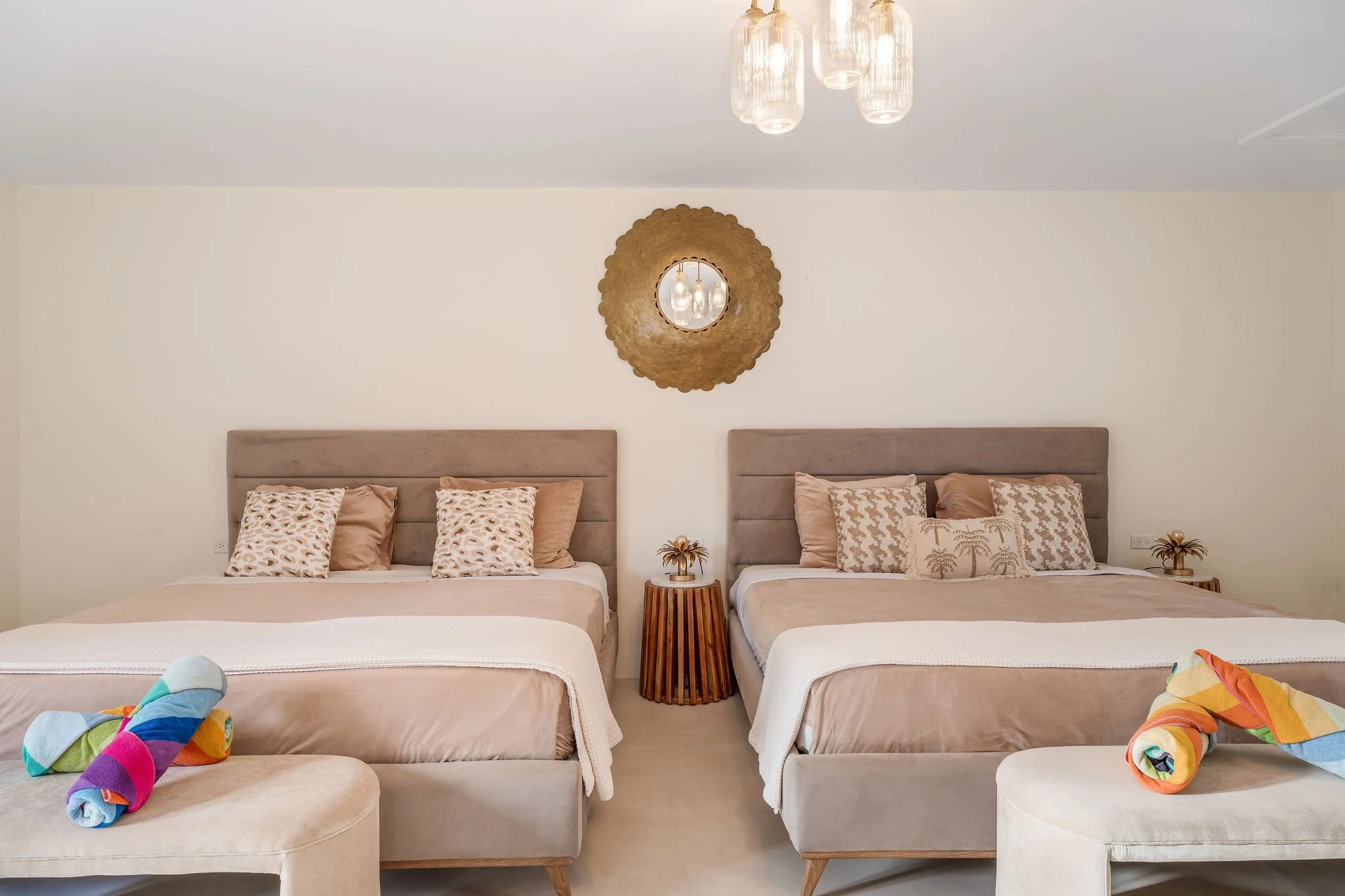 A bedroom with two beds, each with beige headboards, beige bedspreads, and pillows with animal print. Between the beds is a small wooden nightstand with a decorative plant. Above the wall is a circular mirror surrounded by a textured gold frame. Two bedside lamps hang from the ceiling, and near the foot of the beds are colorful rolled-up towels or clothing.