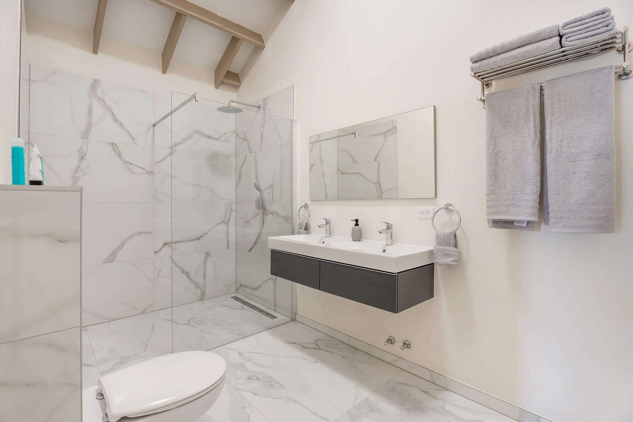 Modern bathroom with marble tiles, glass shower, dual sinks, and gray towels.
