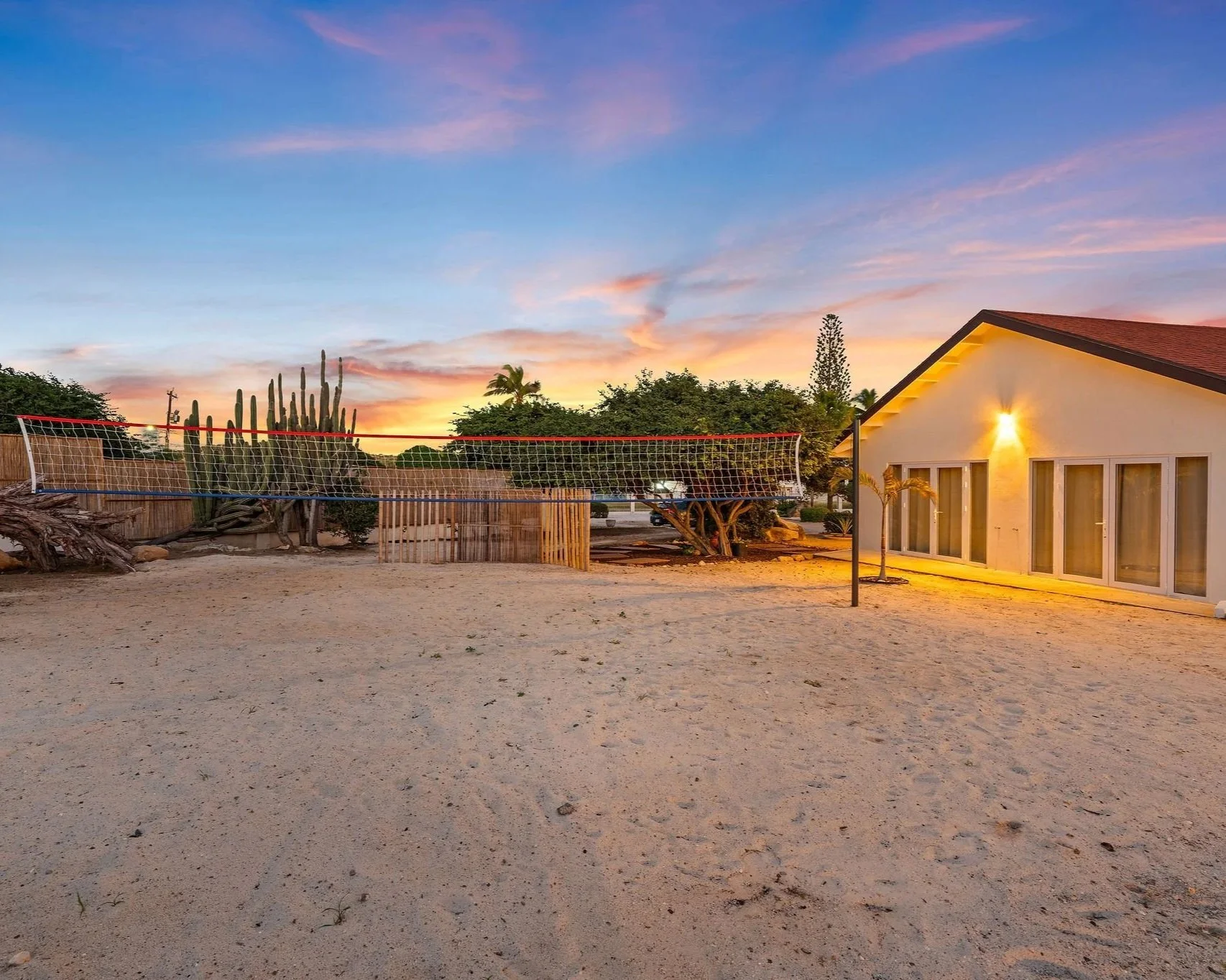 Evening view of a backyard with a volleyball net, desert sand ground, cactus, trees, a house with exterior lighting, and a colorful sky at sunset.