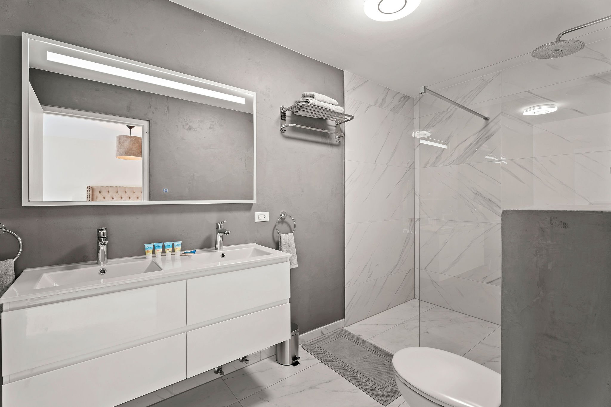 Modern bathroom with a white vanity, large mirror, gray walls, and a walk-in shower with marble tiles. Towels are on a metal shelf, and toiletries are on the sink. Ceiling has recessed lighting and a showerhead is visible.