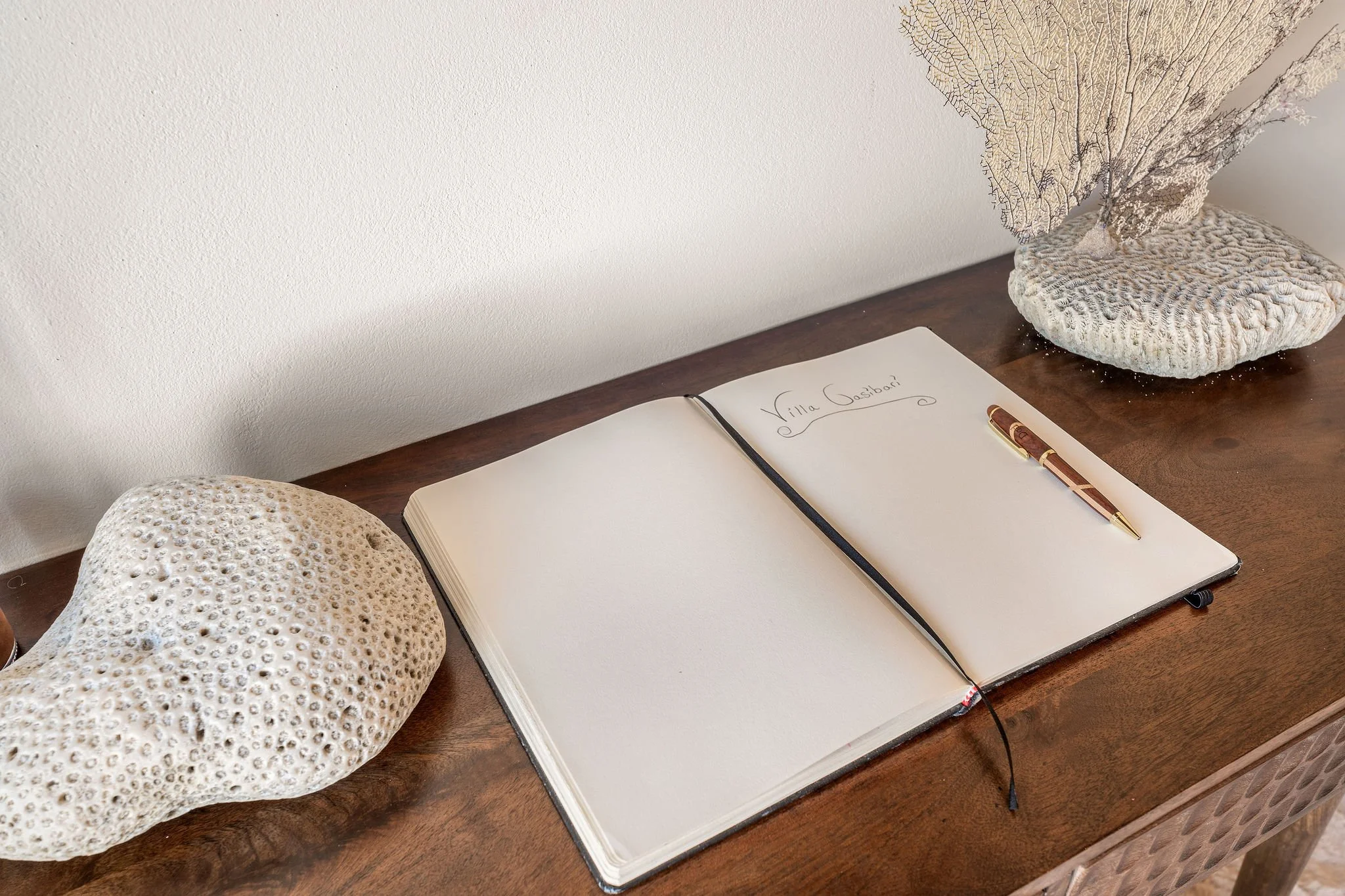 Open notebook with handwritten note saying "Villa Gasbani" on a wooden table, with seashells and a decorative piece resembling coral nearby.