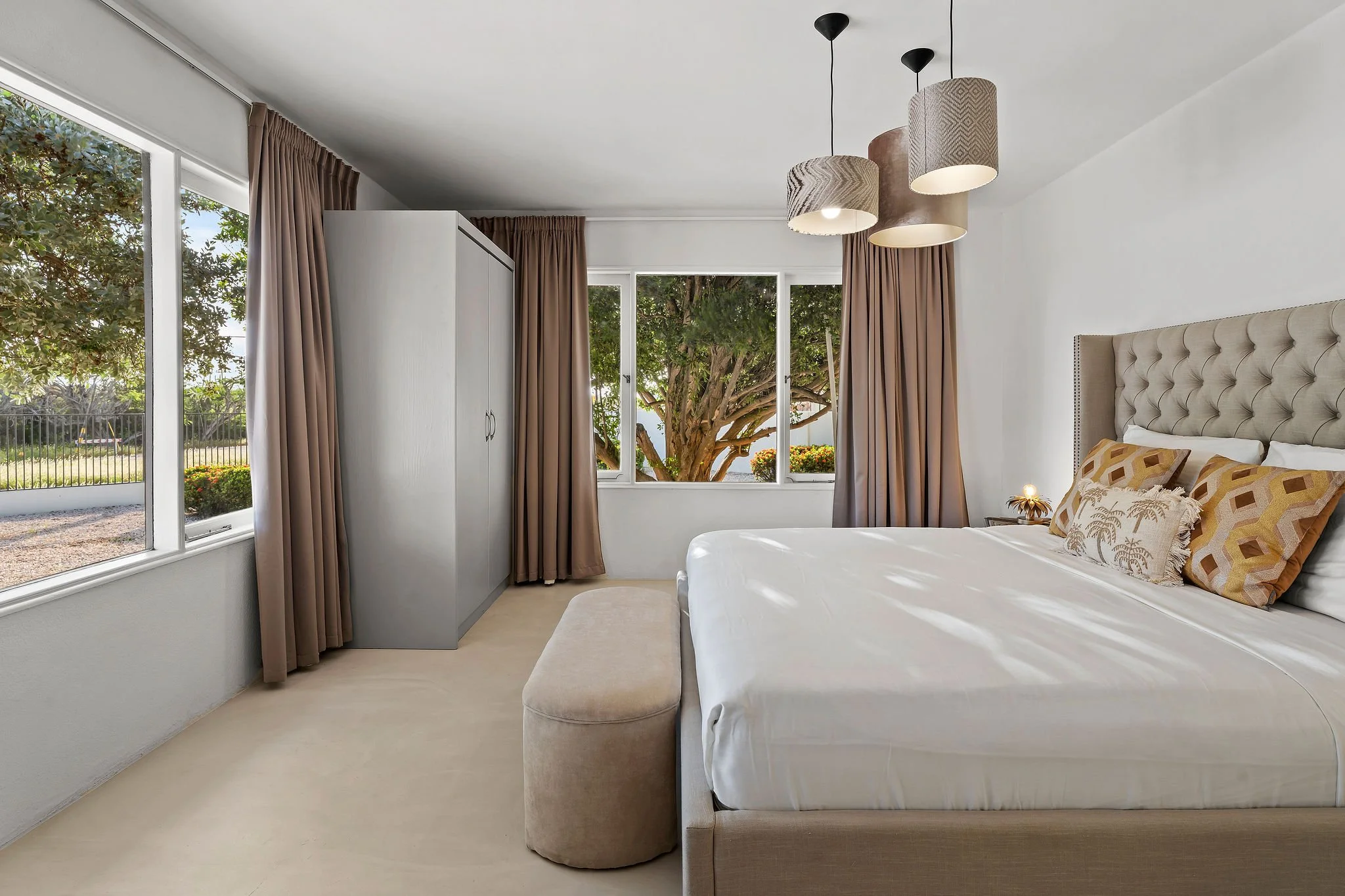 A bright bedroom with large windows, beige curtains, a beige bed with patterned pillows, a white wardrobe, beige ottoman, and modern hanging lamps.