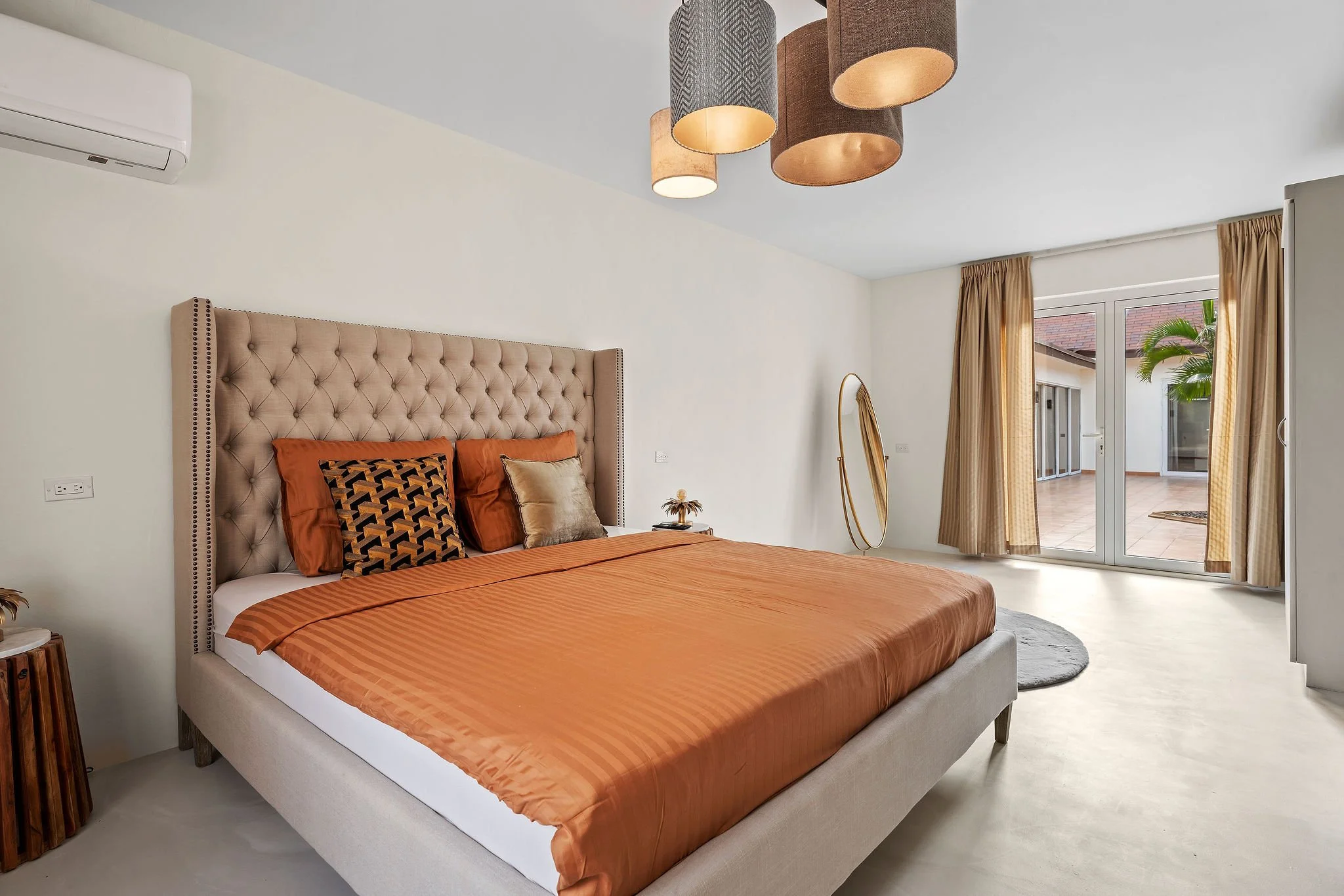 A bedroom with a beige tufted headboard and an orange bedspread with matching pillows. There is a small nightstand with a decorative item, an oval mirror, and a window with beige curtains showing an outdoor balcony.