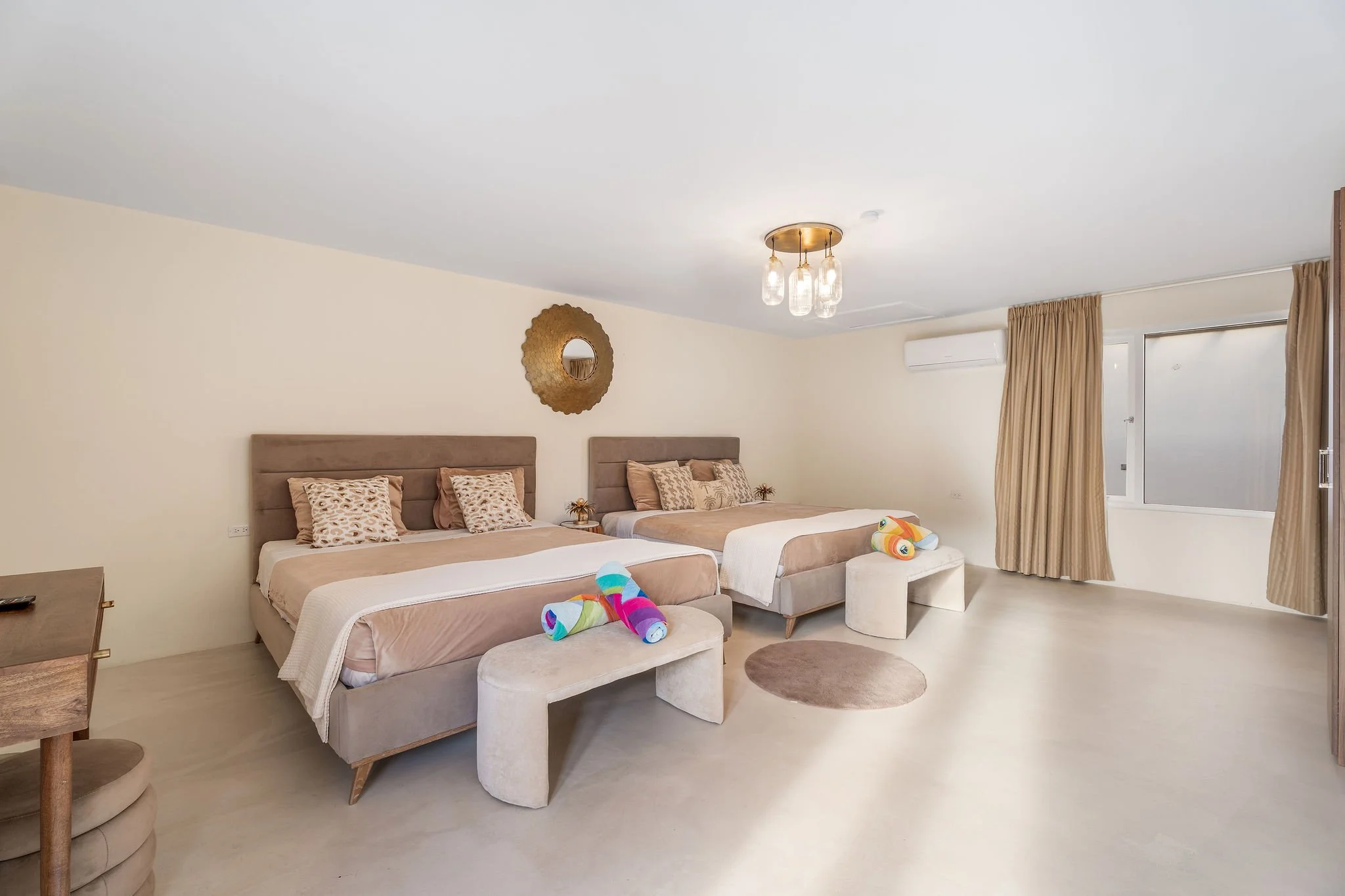 A bedroom with two beds, beige headboards, and patterned pillows. Towels folded into colorful animal shapes are on the foot of each bed. A round mirror hangs on the wall above, and a window with beige curtains is on the right. A light fixture with multiple glass bulbs is on the ceiling.