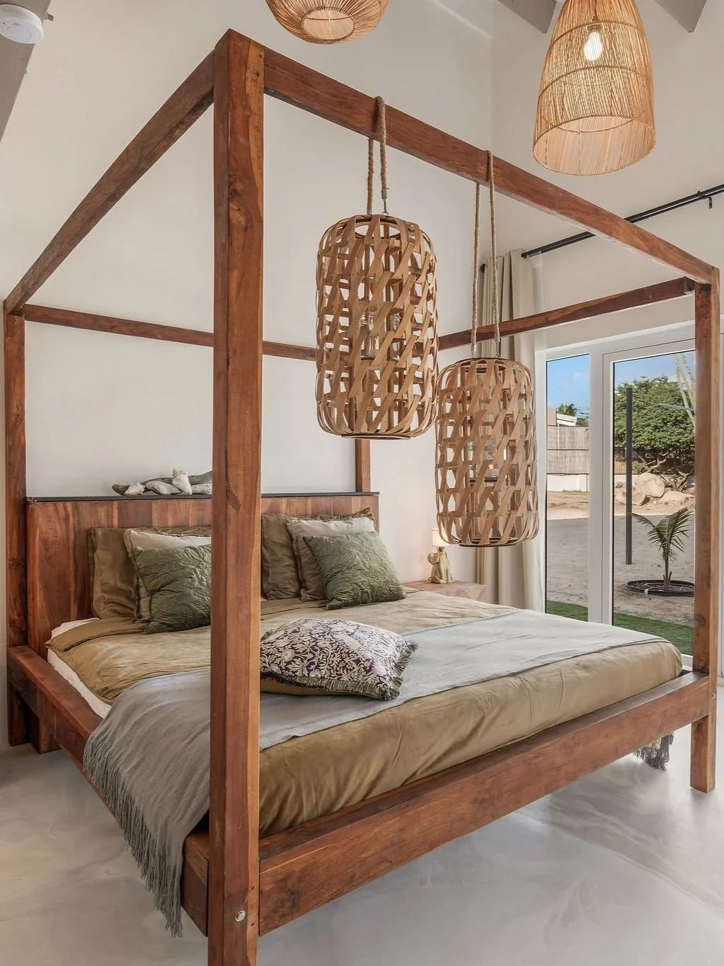 A bedroom with a wooden canopy bed, green pillows, and hanging woven lamps, with large sliding glass doors leading outside to a sandy yard and some plants.