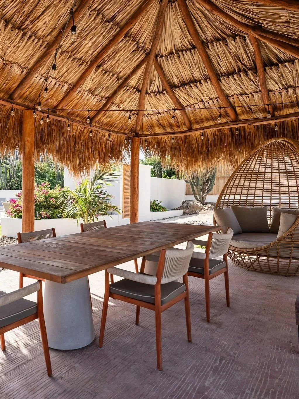 Outdoor patio with a thatched roof, wooden dining table, cushioned chairs, and a hanging wicker egg chair.