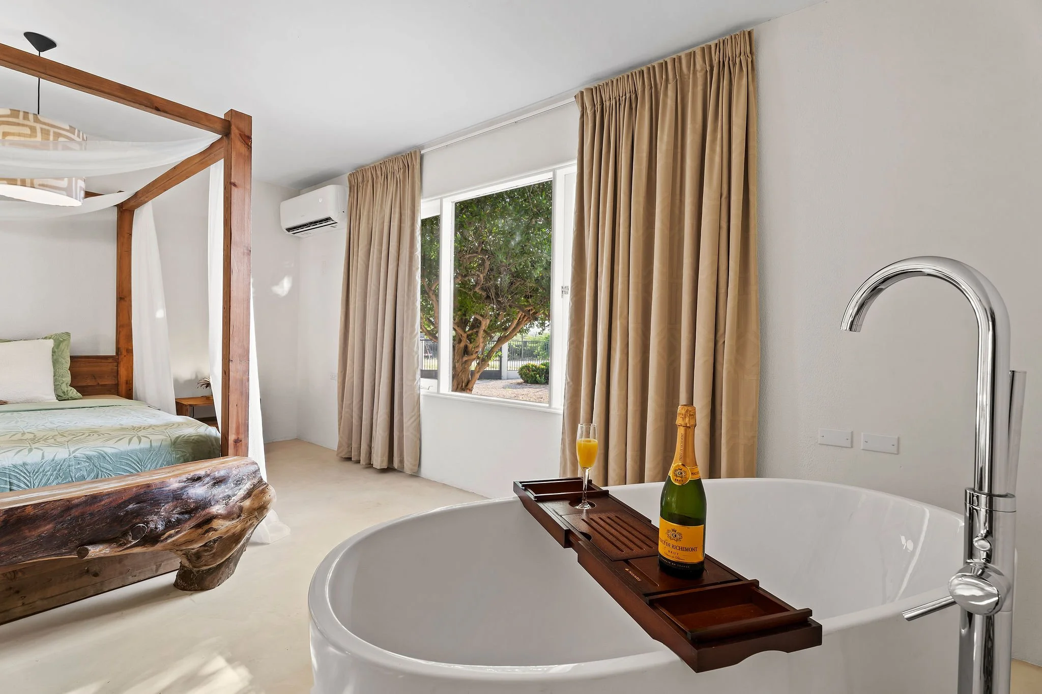 A hotel room with a wooden canopy bed, beige curtains, a window showing trees outside, a white air conditioning unit, a bathtub with a tray holding a bottle of French champagne, a glass of orange juice, and a partially visible sink faucet.
